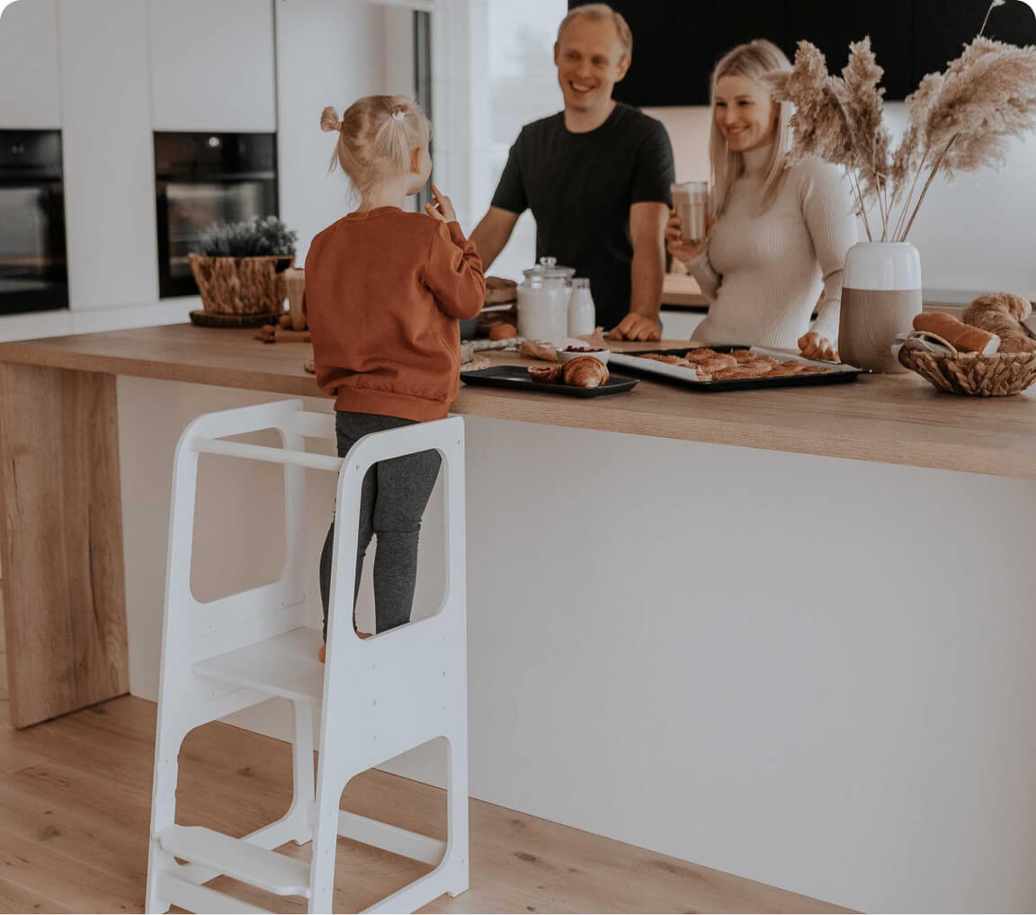 Toddler girl standing on a white kitchen tower in a kitchen cooking with her parents