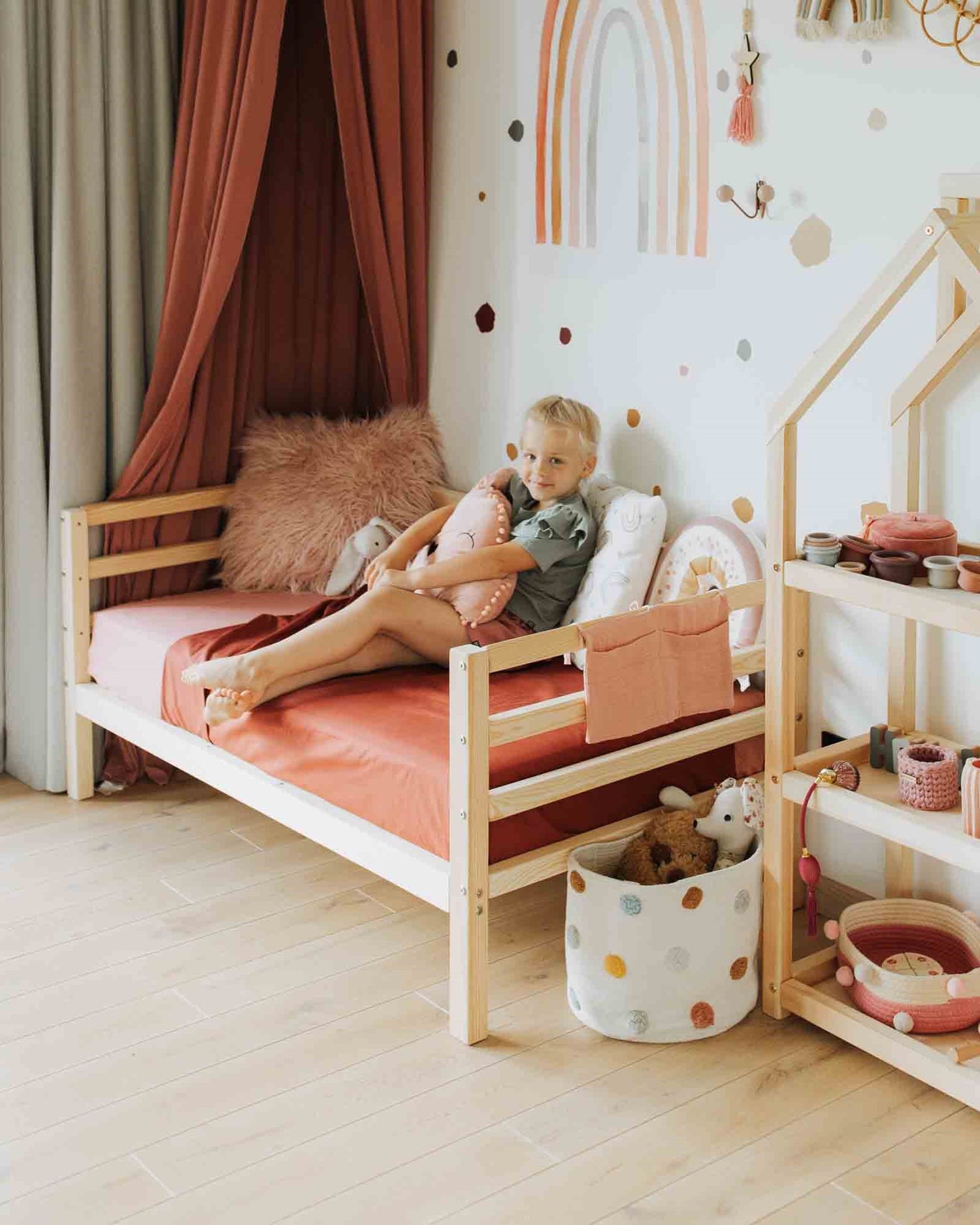 A child sits on a 2-in-1 transformable kids' bed with a 3-sided horizontal rail in a cozy, well-decorated room featuring a rainbow wall decal, wooden furniture with solid wood legs, and various toys. The child's bed and decor are in warm, earthy tones.