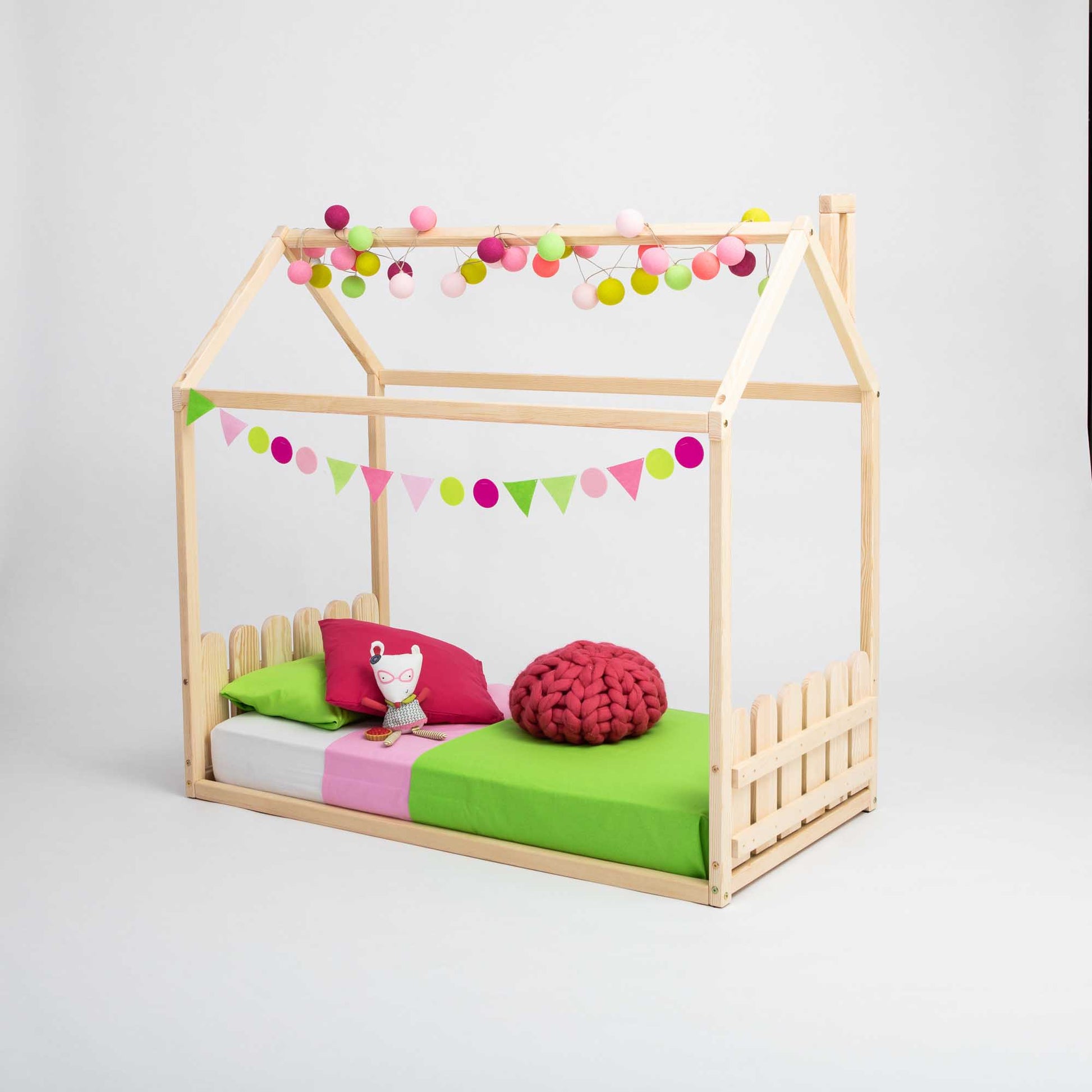Montessori house-frame bed with a picket fence headboard and footboard, decorated with colorful flags and string lights, featuring a green and pink bedding set, a cushion, a knitted red pillow, and complemented by a charming vertical picket fence design.