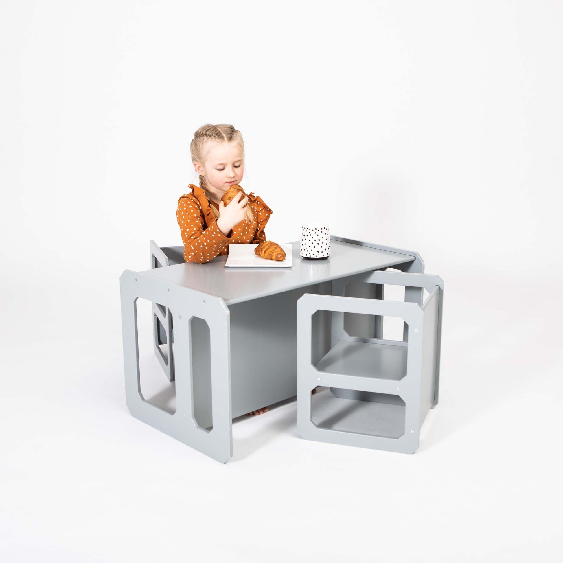 A child sits at a Montessori weaning table and 2 chair set, holding a croissant in one hand and looking at a white polka dot cup, showcasing impressive fine motor skill development.
