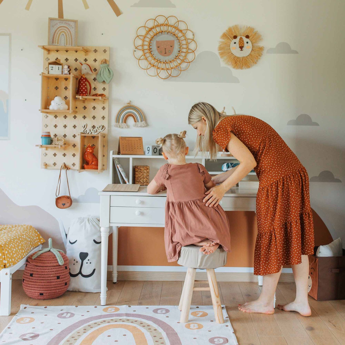 A woman assists a young child seated on a stool at a small desk in a vibrant, decorated room featuring a chic Pegboard Montessori Open Shelf and a rainbow-themed rug, amid various wall hangings.