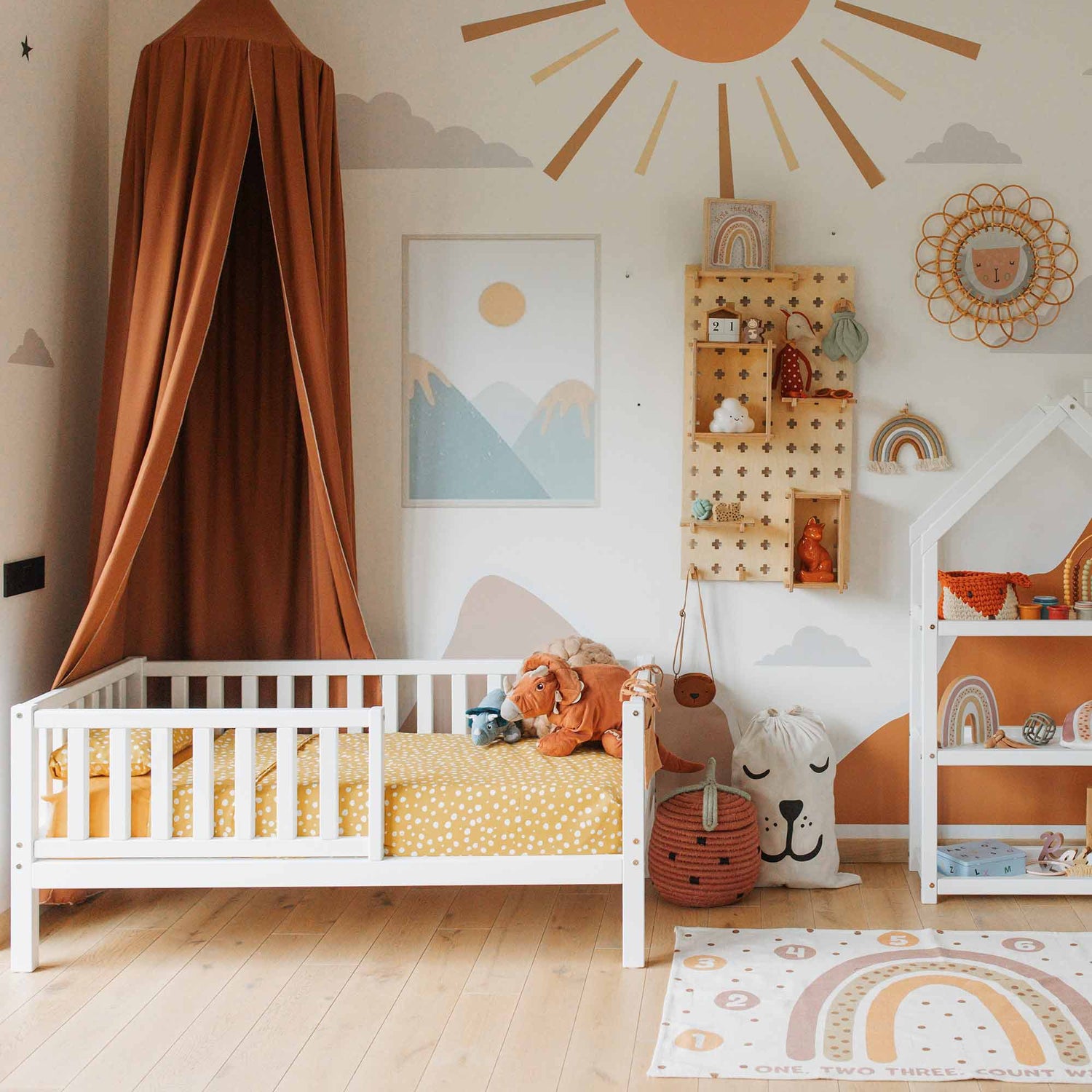 A children's bedroom with a white crib, mustard-colored bedding, and a canopy. The walls are adorned with mountain, sun, and rainbow illustrations. The Pegboard Montessori Open Shelf offers a clever storage solution for toys and decor items. The setup creates a cozy Sweet Home from Wood ambiance.