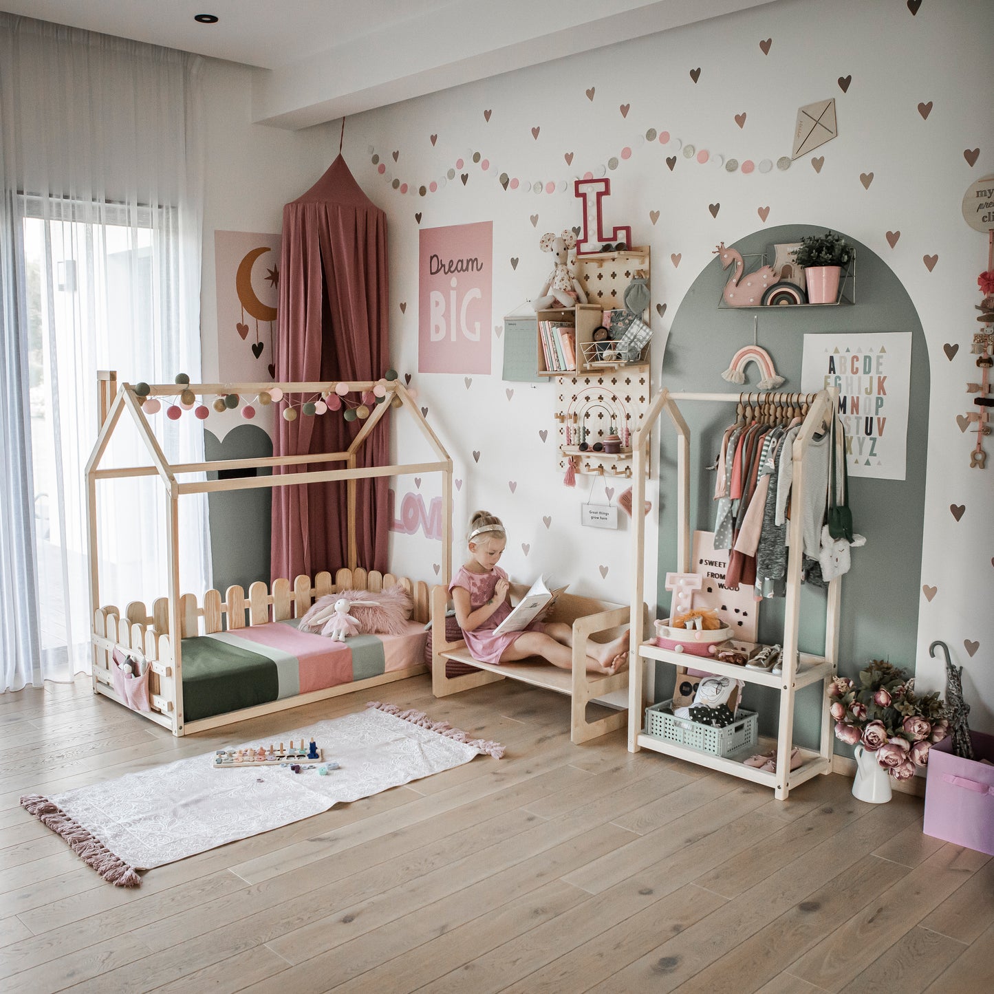 A young girl reads a book in a decorated children's bedroom featuring a Floor house floor bed with 3-sided picket fence rails, an open closet, toys, and playful wall art.