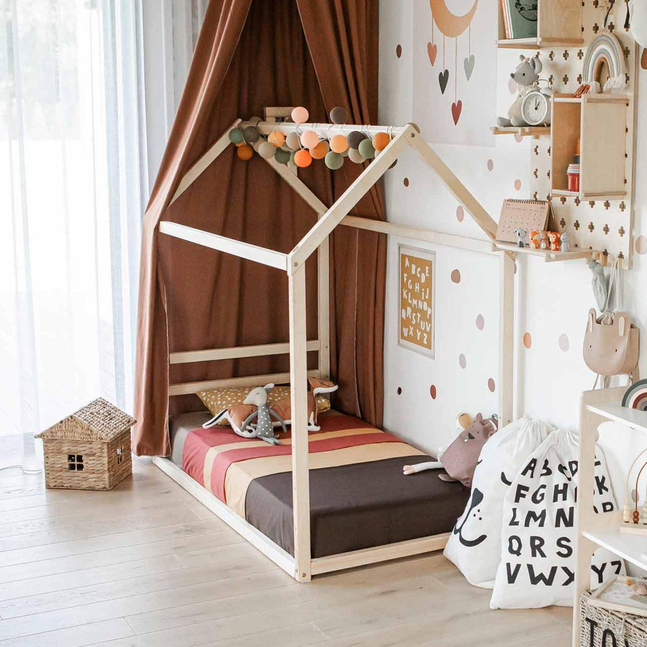 A cozy children's bedroom features a Children's house bed with a horizontal headboard adorned with soft toys. Shelves brimming with books and toys, and a letter-patterned wall hanging add charm. Light wood flooring and a window draped in sheer curtains complete the inviting space.