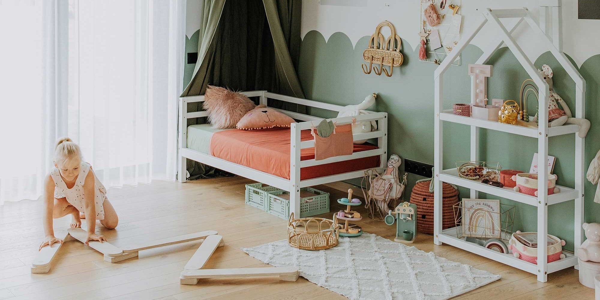 A child playing with wooden blocks on the floor in a well-organized, pastel-themed bedroom featuring a 2-in-1 transformable kids' bed with solid wood legs and a 3-sided horizontal rail, along with toys and shelves.