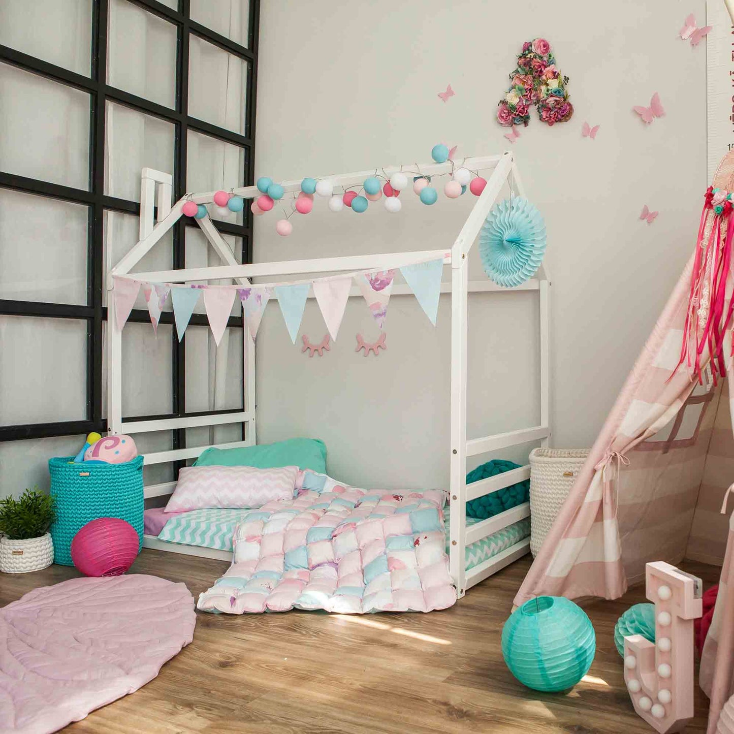 A children's room featuring a Floor house-frame bed with a horizontal headboard and footboard, adorned with pastel-colored bunting, string lights, and cozy bedding. The room is filled with various toys and decor items, creating a playful atmosphere.