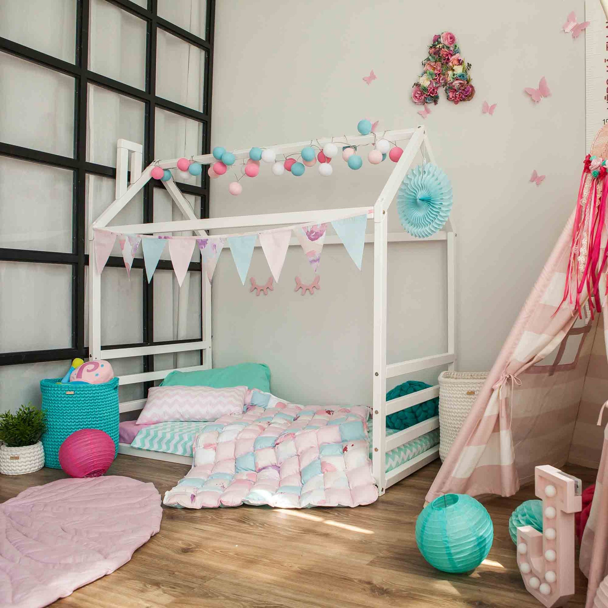 A children's room featuring a Floor house-frame bed with a horizontal headboard and footboard, adorned with pastel-colored bunting, string lights, and cozy bedding. The room is filled with various toys and decor items, creating a playful atmosphere.