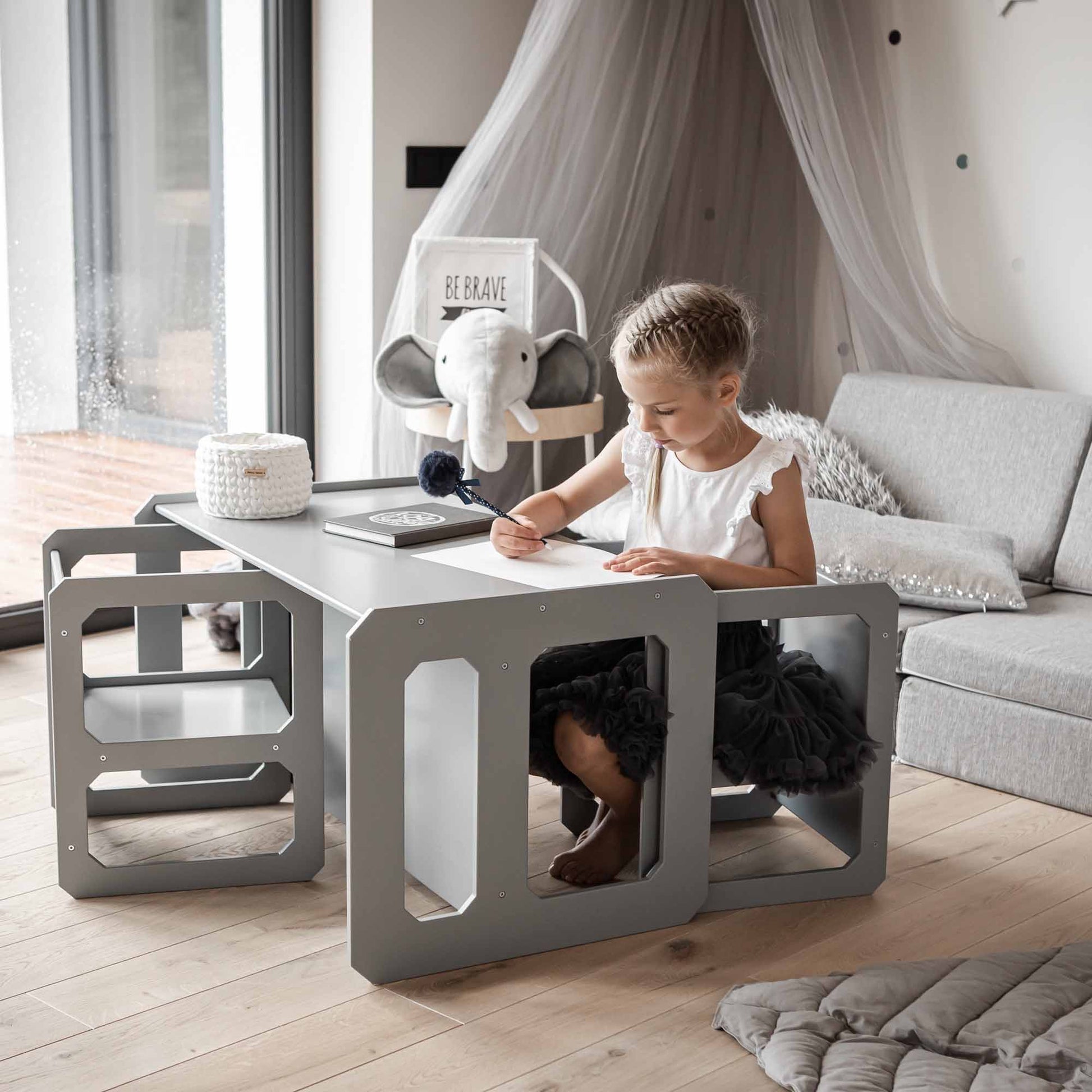 A young girl sits at a Montessori weaning table set with two chairs, designed in modern, multi-functional grey with built-in shelves, fostering fine motor skill development as she draws on a piece of paper. The room is decorated with neutral tones and an elephant headpiece on the back of the chair.