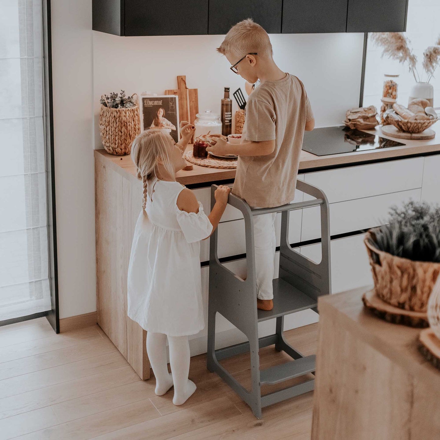 Two toddlers standing on a 2-in-1 Convertible kitchen tower - table and chair in a kitchen.