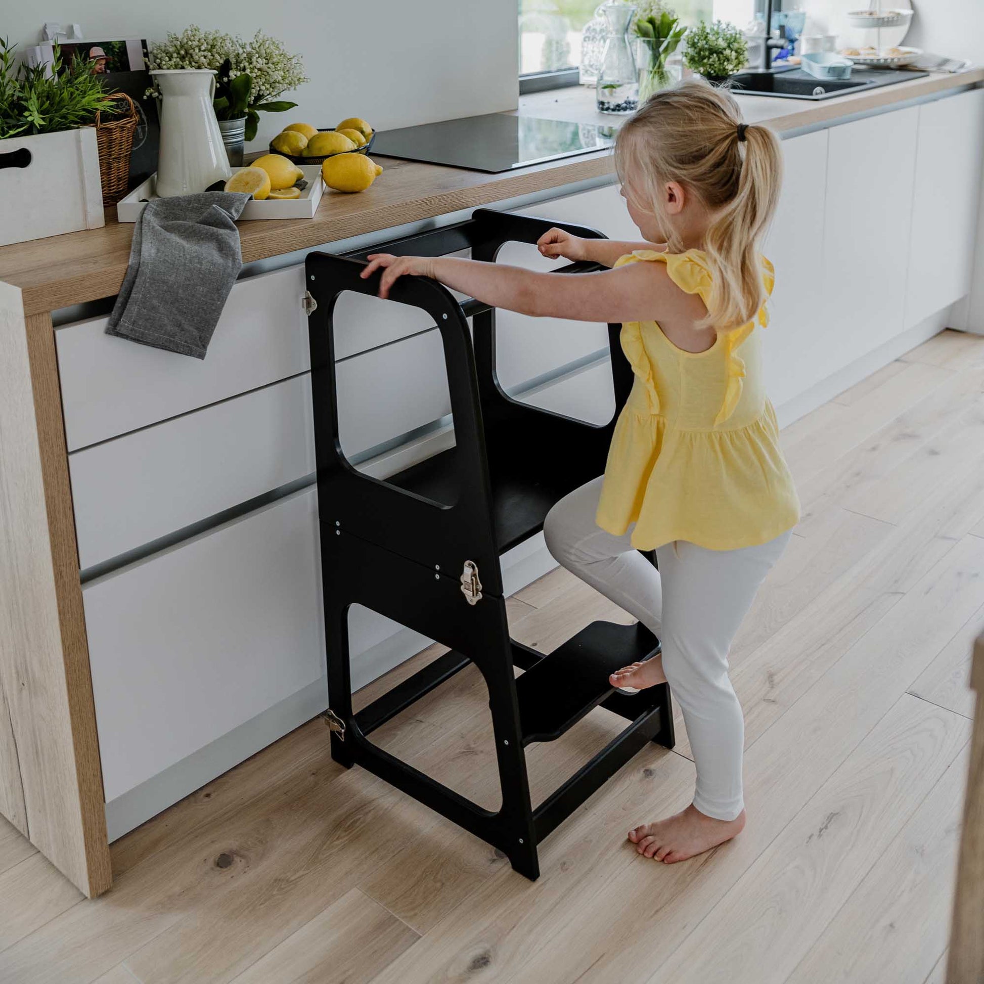 A toddler standing on a 2-in-1 Convertible kitchen tower - table and chair in a kitchen.