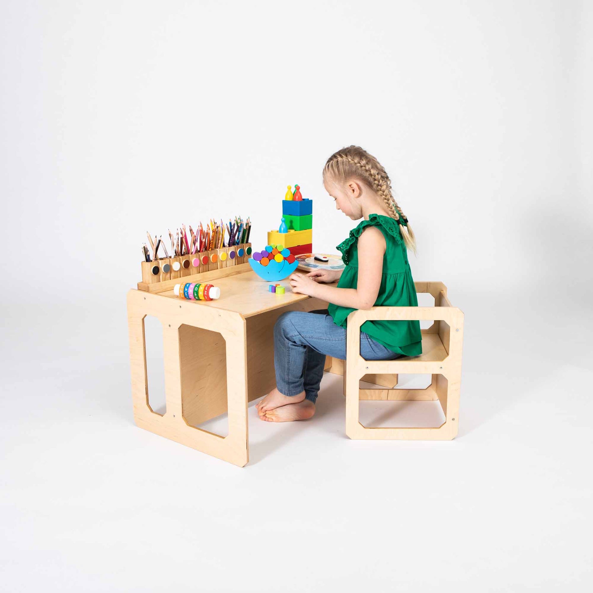 A child in a green shirt and blue jeans sits at a Montessori weaning table and chair set, playing with colorful building blocks. Various art supplies are arranged on the desk, creating a perfect Montessori-inspired environment that encourages toddler independence and fine motor skill development.