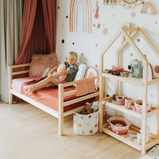 A child sits on a Kids' bed on legs with horizontal rail on 3-sides, topped with pink bedding in a decorated bedroom featuring a toy house shelf, plush toys, and rainbow and dot-themed wall art.
