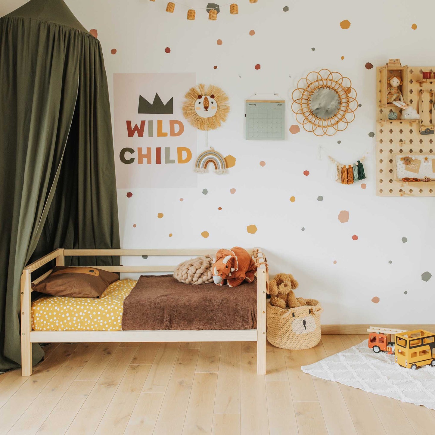 Children’s bedroom featuring a kids' bed on legs with horizontal rail on 3 sides, canopy, wall decor, toys, basket with stuffed animals, and colorful polka dot wall stickers.