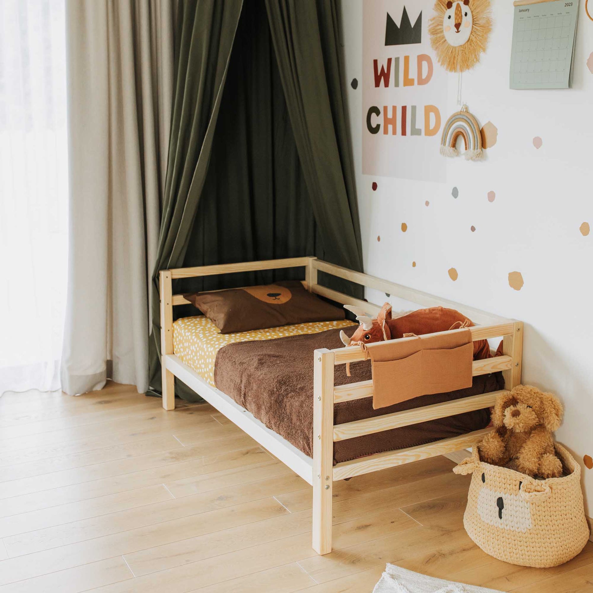 A Kids' bed on legs with a horizontal rail on 3 sides, featuring brown bedding, sits beneath a green canopy in a decorated room with stuffed toys, a basket, and "WILD CHILD" lion wall art.