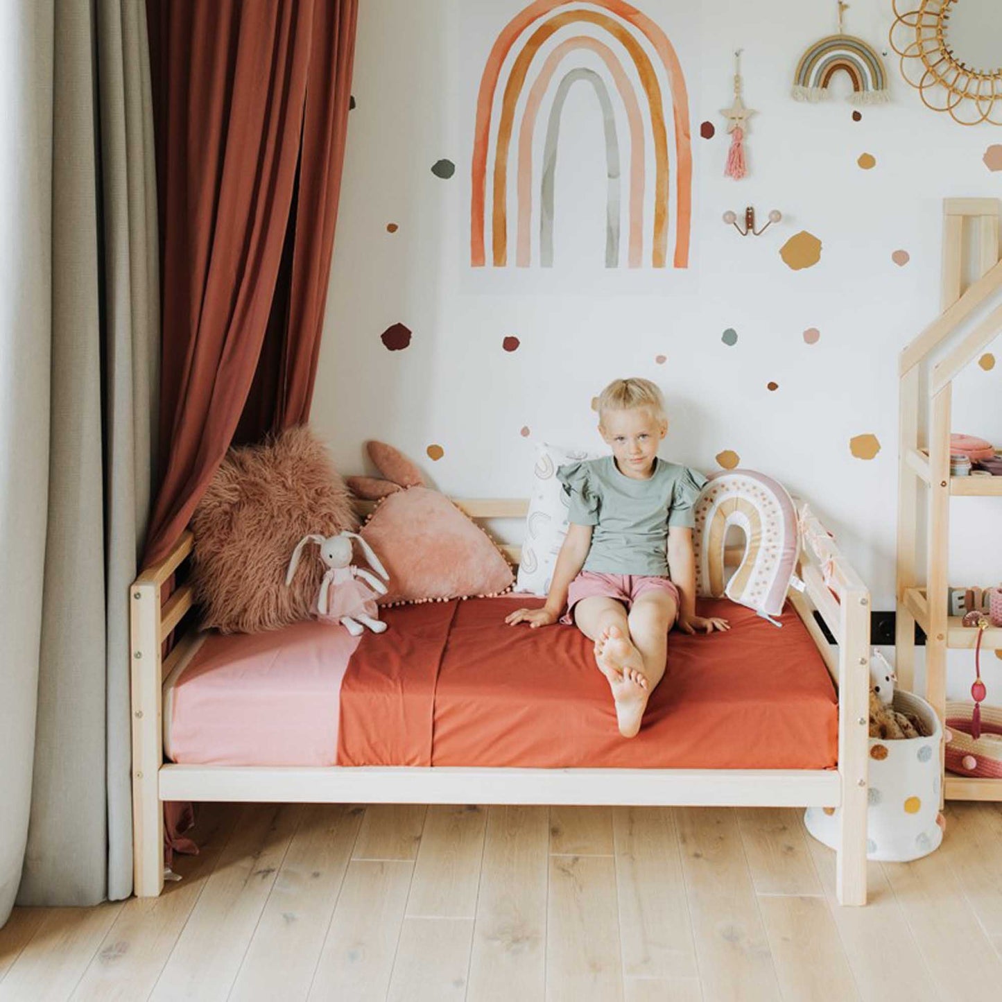 A young child sits on a Kids' bed on legs with horizontal rail on 3-sides, featuring red bedding in a rainbow-themed bedroom filled with soft toys.