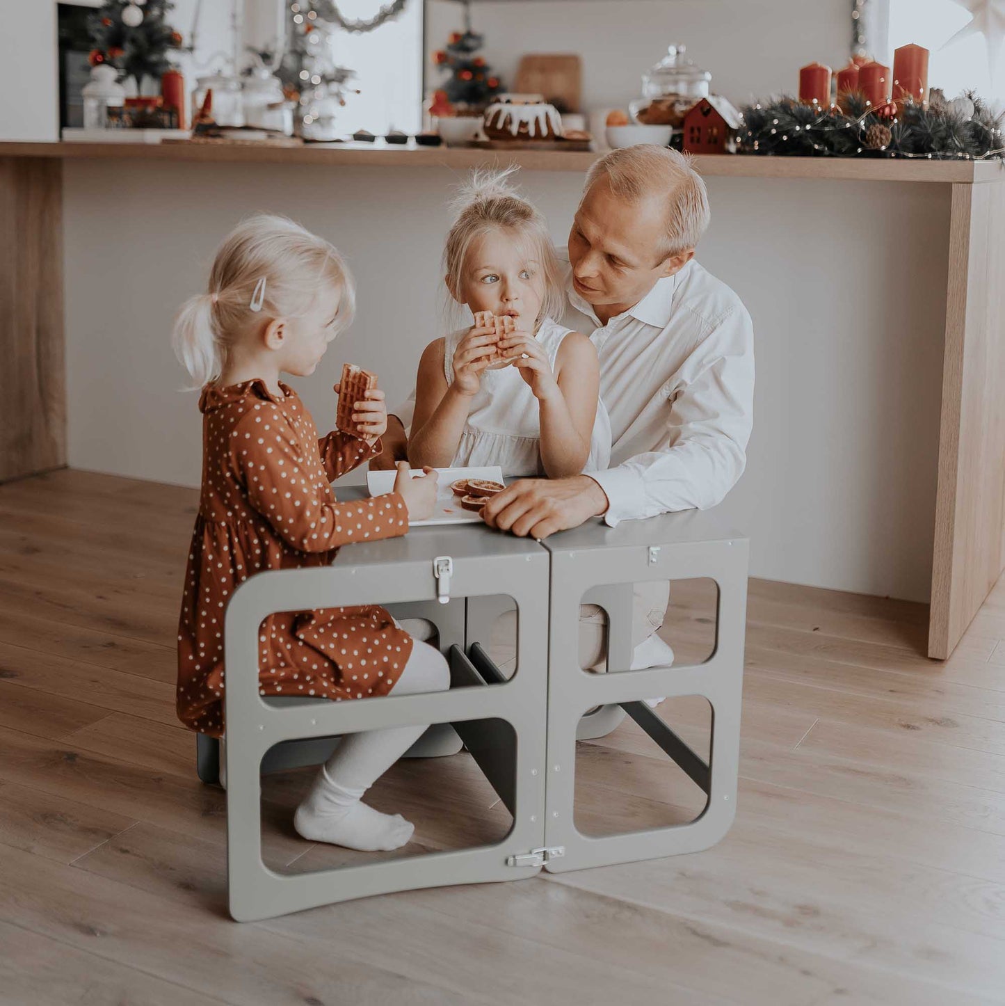 A man and two children sitting at a table in a kitchen with a Sweet Home From Wood 2-in-1 transformable kitchen tower - table and chair set.