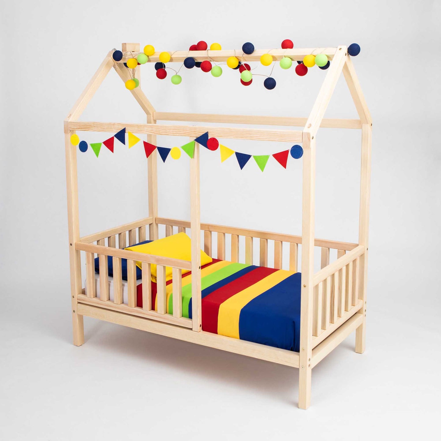 The Kids' house bed on legs with a fence features a wooden frame shaped like a house, complete with roof-like beams. It is elegantly decorated with colorful hanging balls and triangular pennant banners, and comes with a vibrant striped mattress and pillow set—ideal for Montessori-style co-sleeping arrangements.