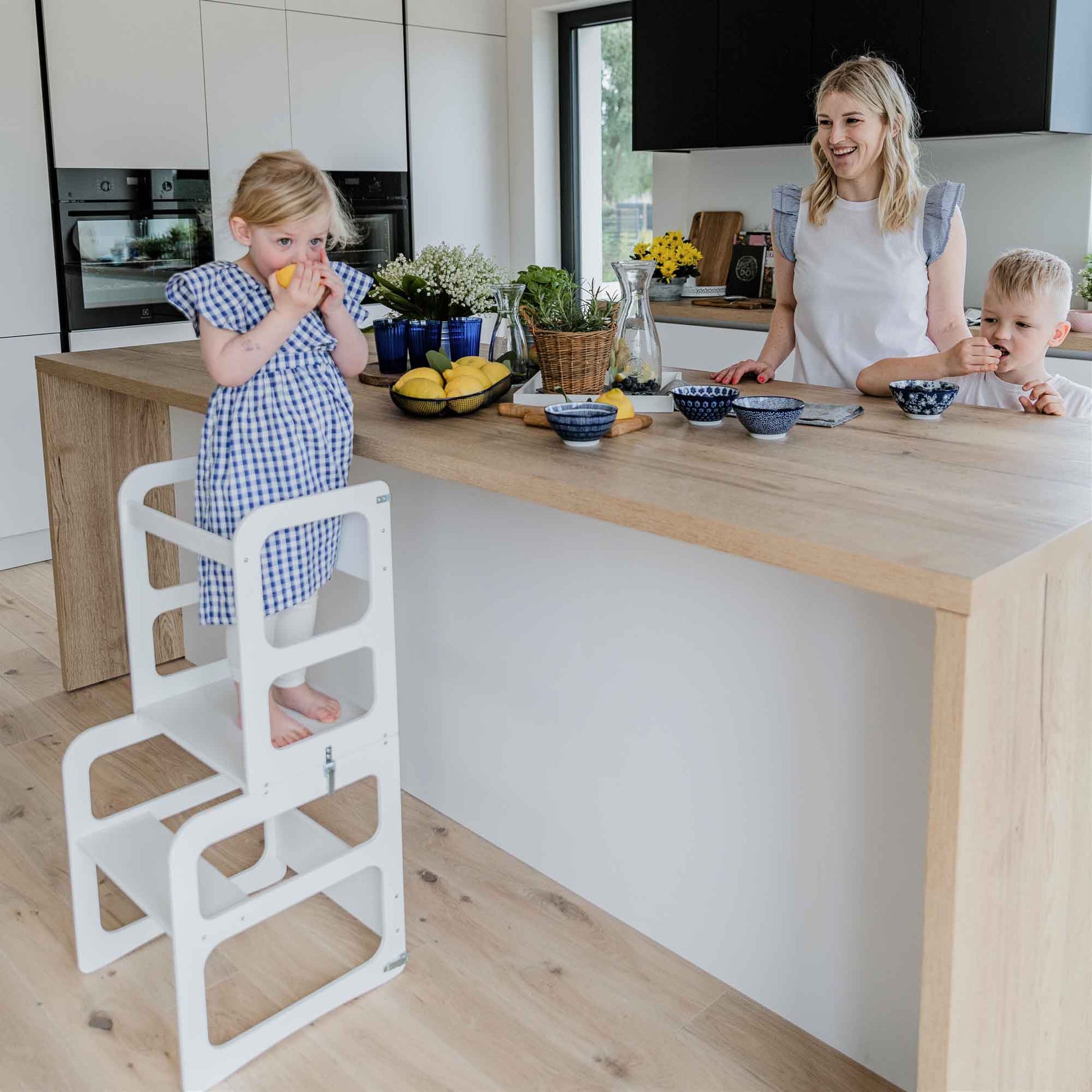 A woman stands in a modern kitchen smiling while a child on a step stool, the 2-in-1 transformable kitchen tower - table and chair set, eats an orange. Another young chef stands beside the counter. Various kitchen items and fruits are visible on the counter.