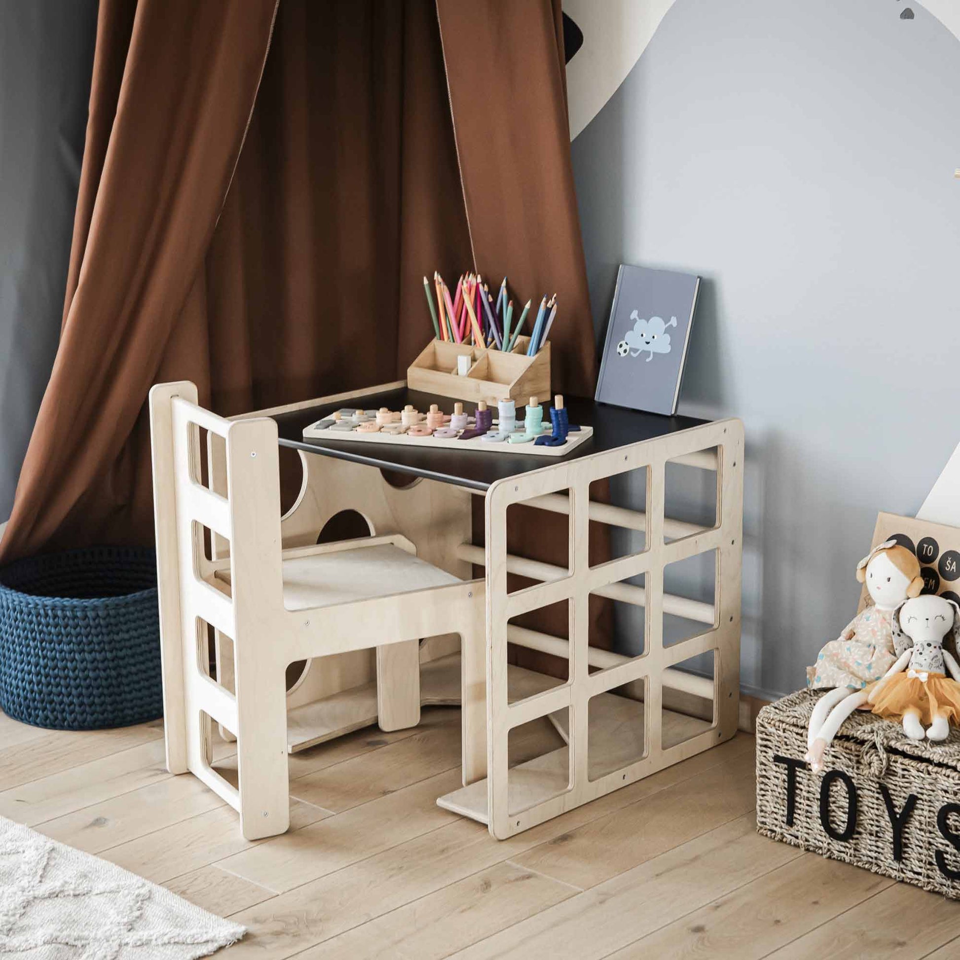 A small wooden child's desk with a chair, paint palette, colored pencils, and a chalkboard sits next to a Montessori table in a room featuring a brown tent and hardwood flooring. Nearby, there's a basket of toddler toys and even a 2-in-1 table and chair set or activity cube for endless adventures.