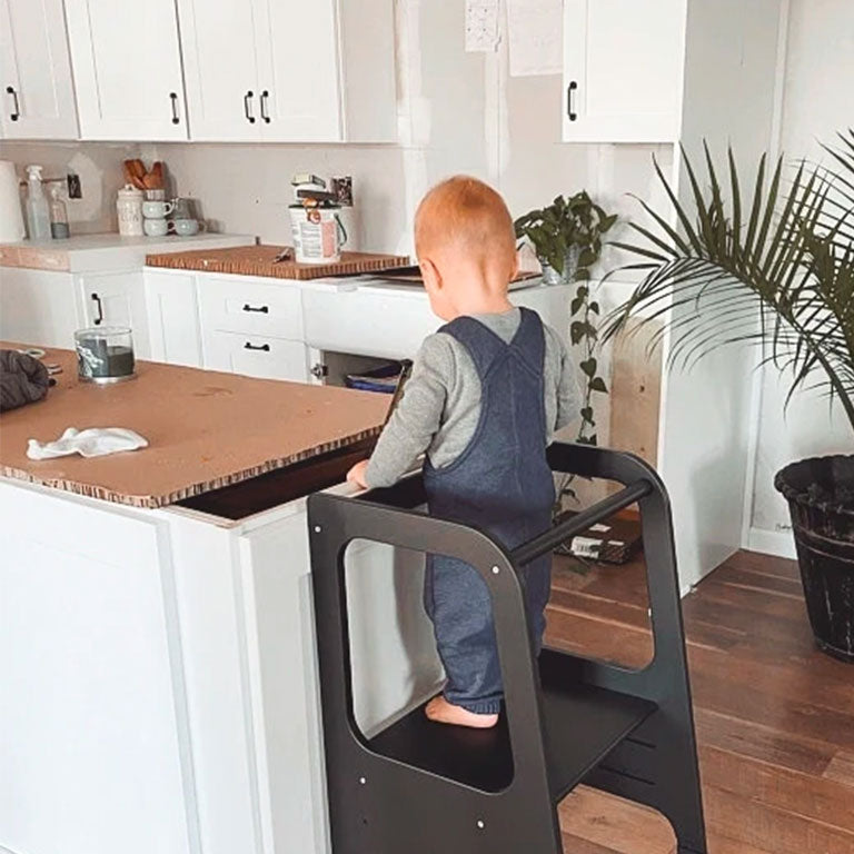 Customer photo of a baby boy standing on a toddler step stoll in a kitchen.