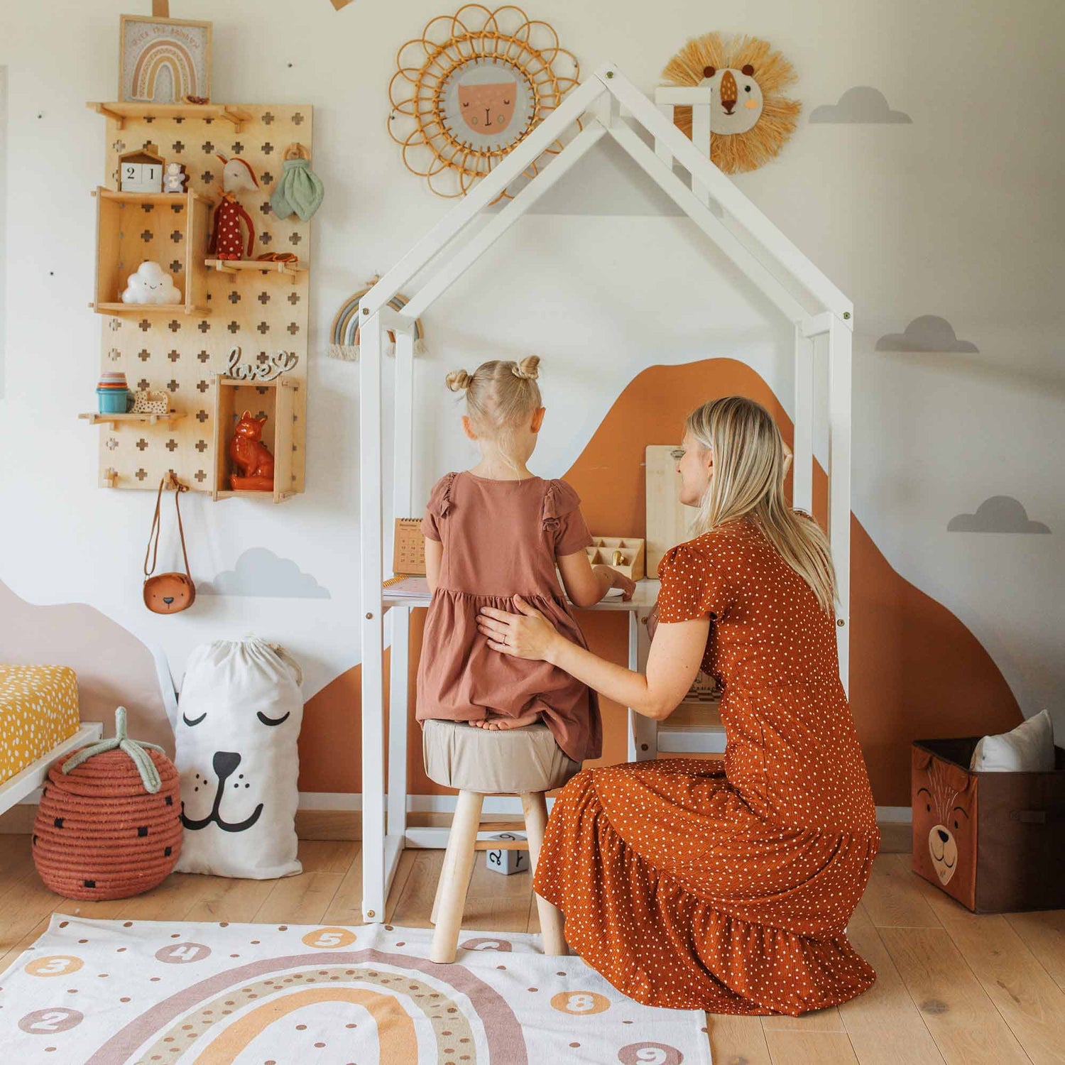 A woman in an orange dress kneels and comforts a pre-schooler sitting at a House-shaped Toddler Desk in a playroom. The room features shelves filled with toys, wall decorations, and a white toy storage bag with a face design.