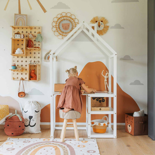 A pre-schooler in a brown dress sits at the House-shaped toddler desk in a room decorated with wall shelves, baskets, and animal-themed items. The wall features a mountain mural and cloud stickers, perfectly accentuating the built-in storage.