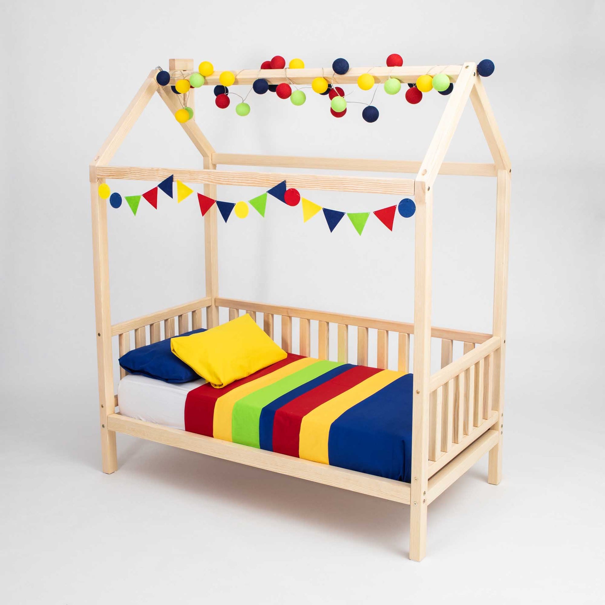 A Raised house bed on legs with 3-sided rails, featuring a multicolored striped bedspread, yellow pillow, and decorative string lights along with triangular flags hanging from the frame. This cozy setup is perfect for co-sleeping families who value a vibrant yet comforting bedtime environment.