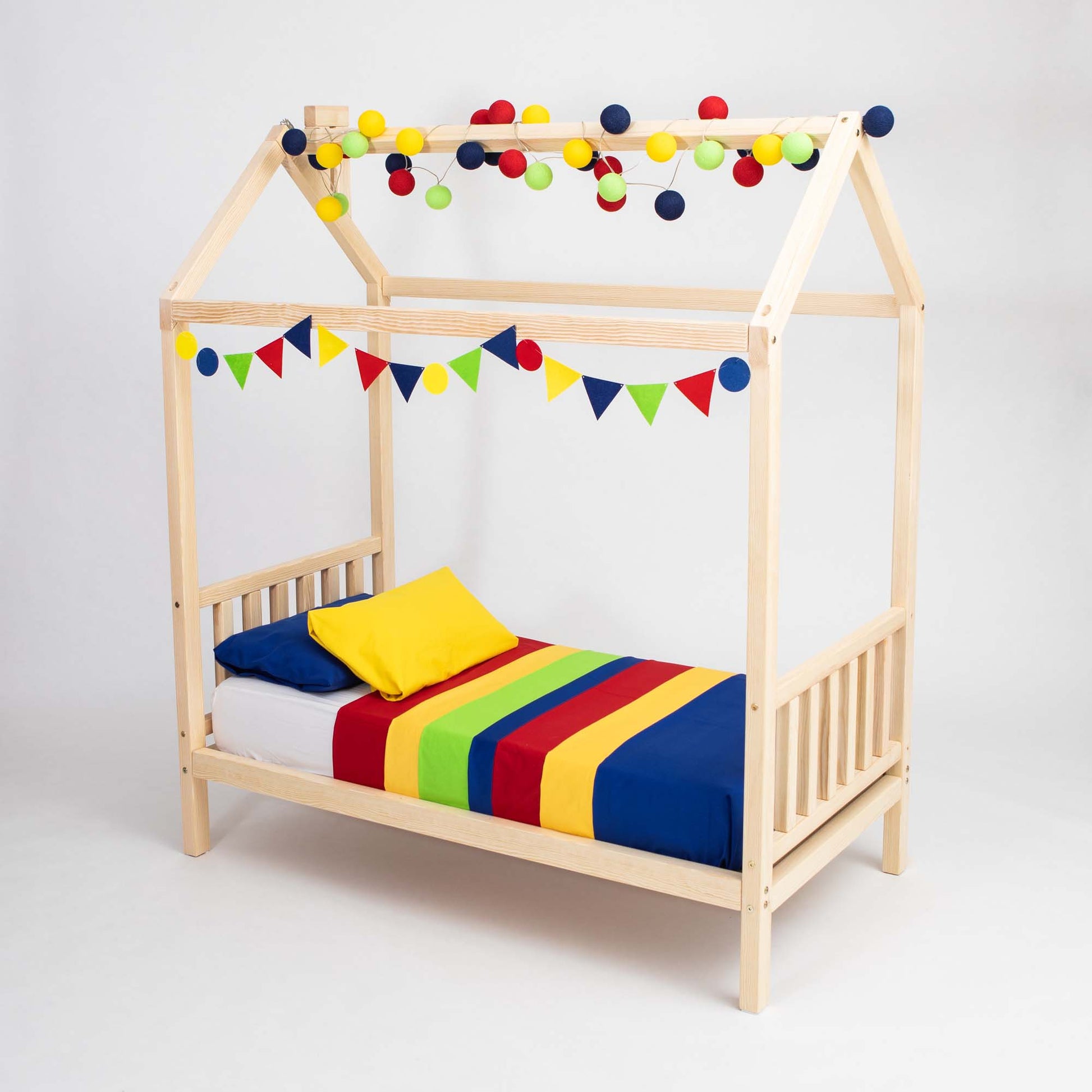 The Toddler House Bed on Legs with a Headboard and Footboard is a charming wooden bed shaped like a house. It is decorated with vibrant pom-poms and triangular flags, complete with a rainbow-striped blanket and a cheerful yellow pillow, making it the ideal sleeping space for kids.