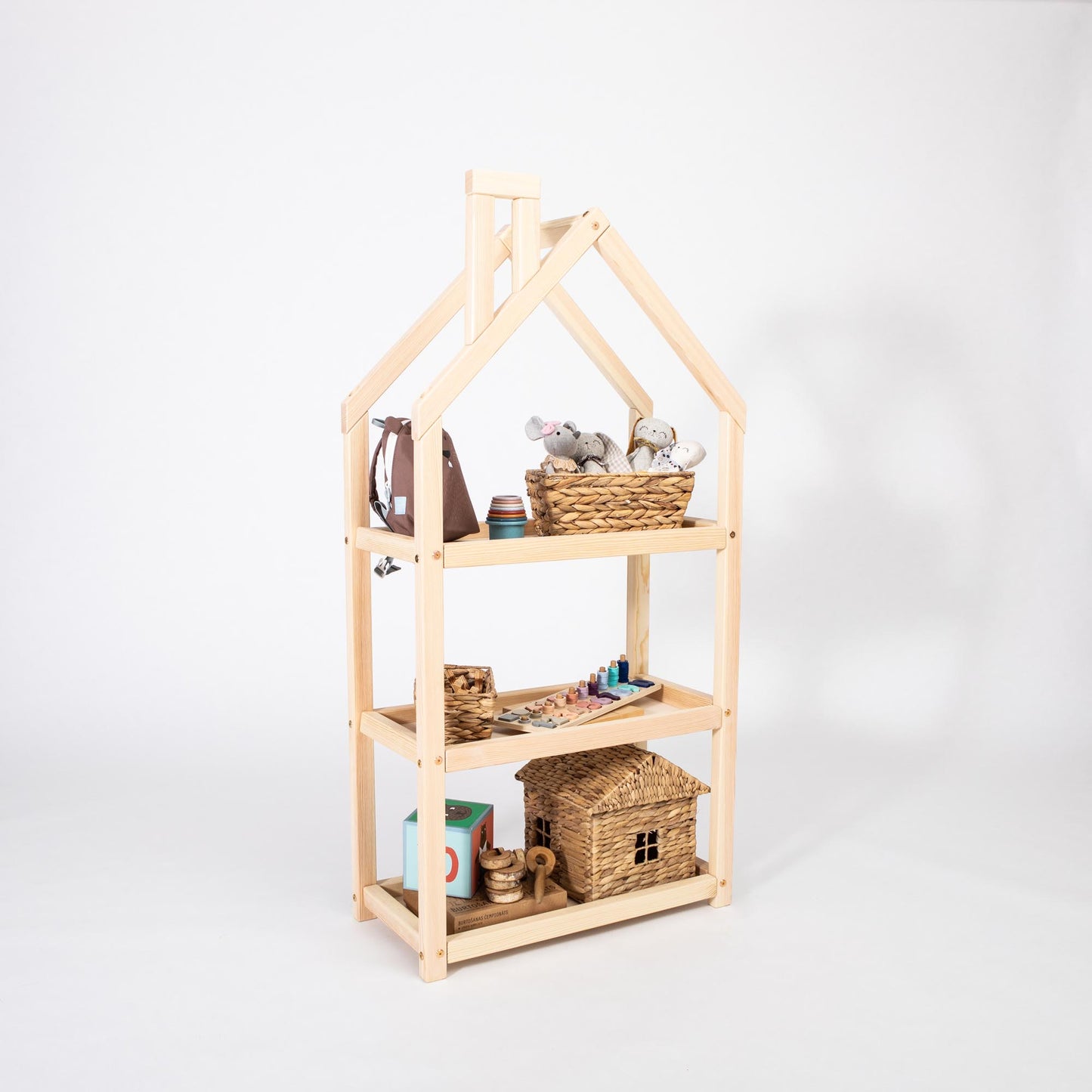 The House-shaped Montessori shelf is a minimalist, three-tiered wooden piece that beautifully displays various items like stuffed animals, baskets, a toy house, and art supplies. Ideal as nursery shelves, this Montessori-inspired design keeps your space organized and stylish.