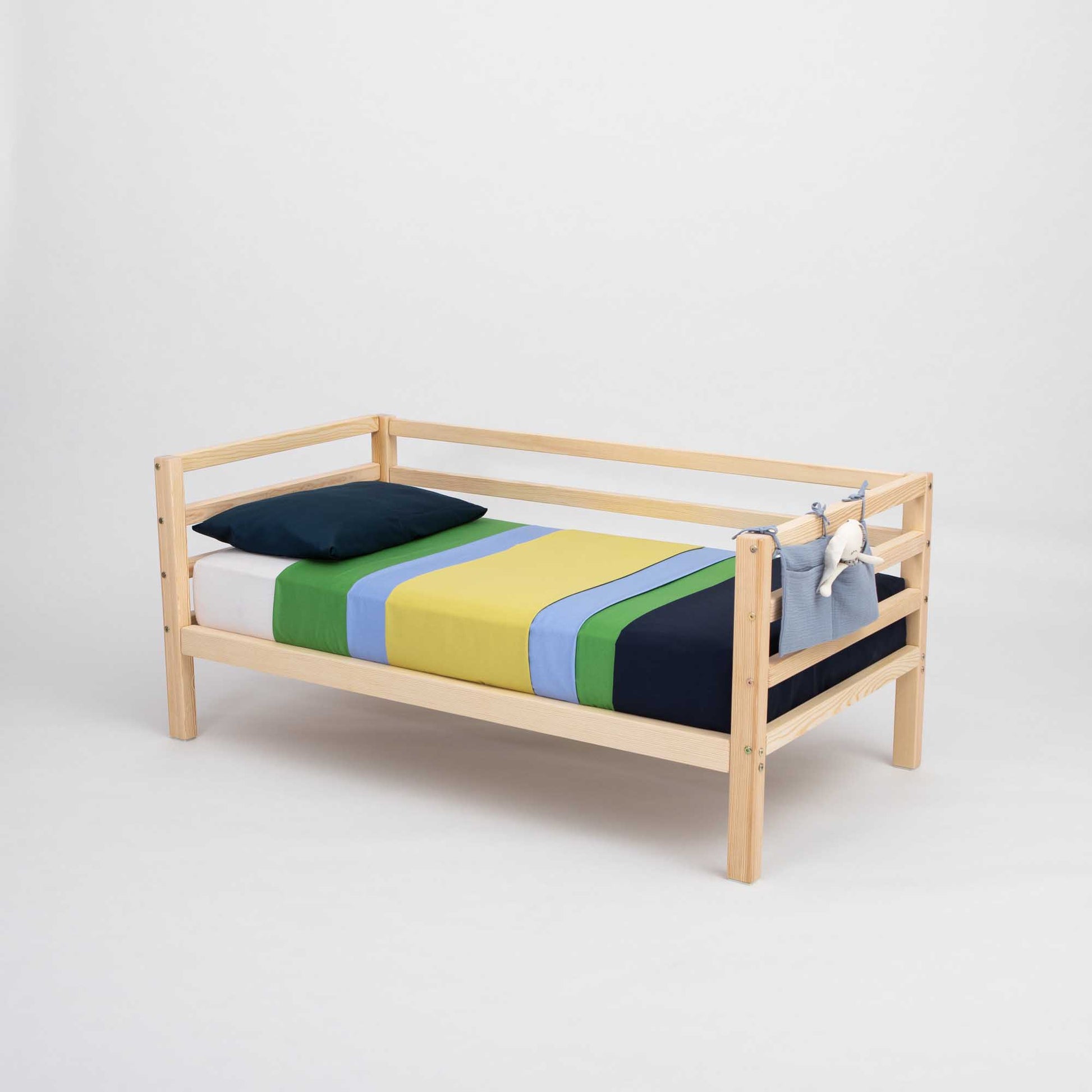 A solid wood Kids' bed on legs with horizontal rail on 3-sides, featuring striped blue, green, yellow, and white bedding, a dark blue pillow for comfort, and stuffed toys hanging from the side for added fun.