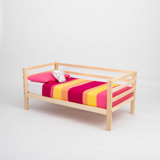 The Kids' bed on legs with a horizontal rail on 3 sides includes a mattress, a cartoon character pillow, and striped bedding in red, orange, pink, and yellow tones. Crafted from solid wood and inspired by Montessori design for toddlers.