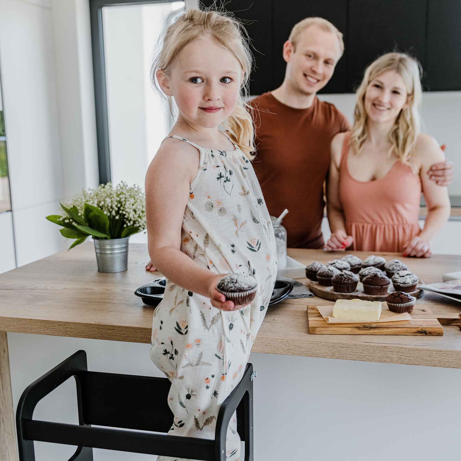 A young chef holding a cupcake stands on a 2-in-1 transformable kitchen tower - table and chair set by a kitchen counter, with her parents smiling in the background. The counter is adorned with more cupcakes and a plant.