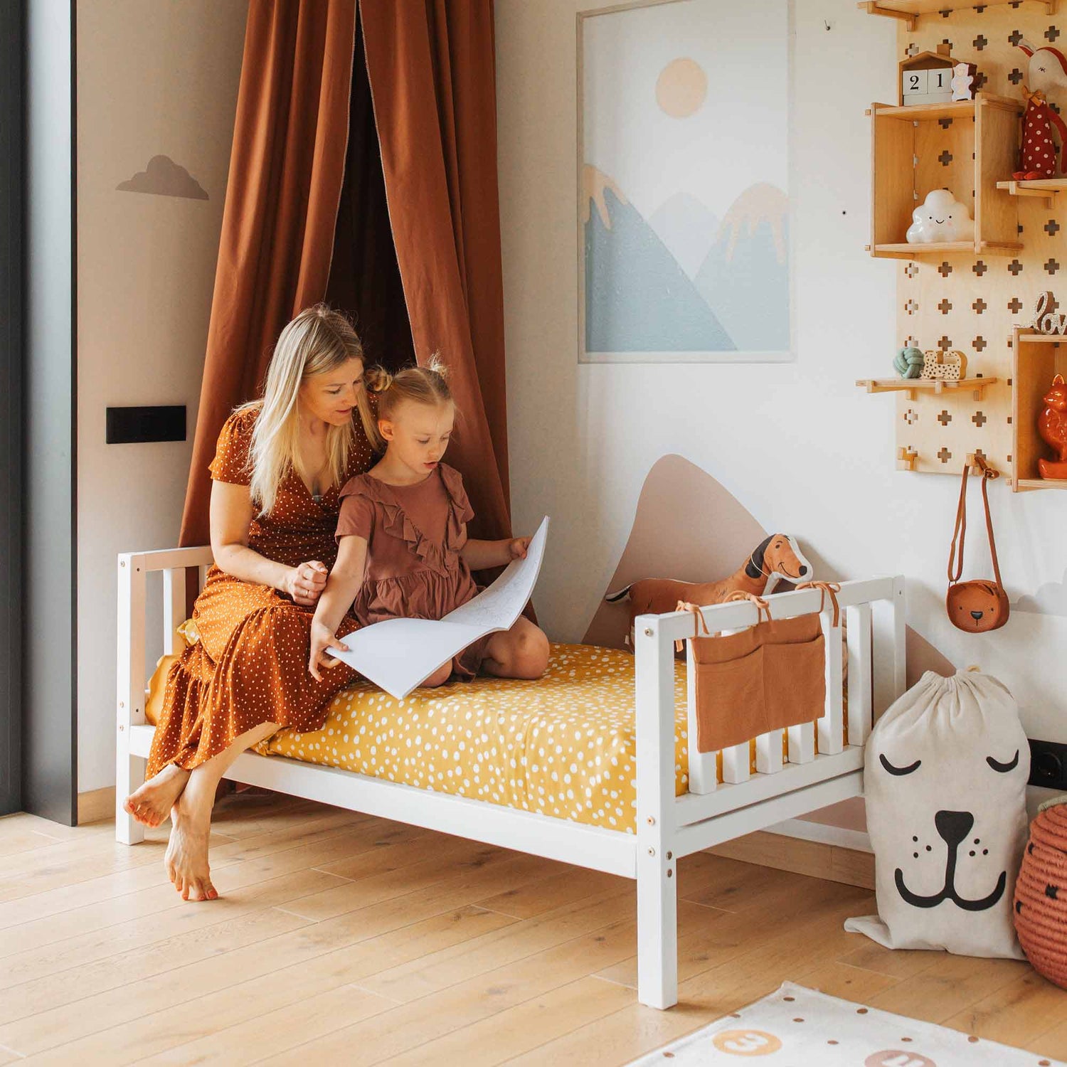 A woman and a child sit on a Raised kids' bed on legs with a headboard and footboard in a tidy, well-decorated room; the woman is reading a book to the child.