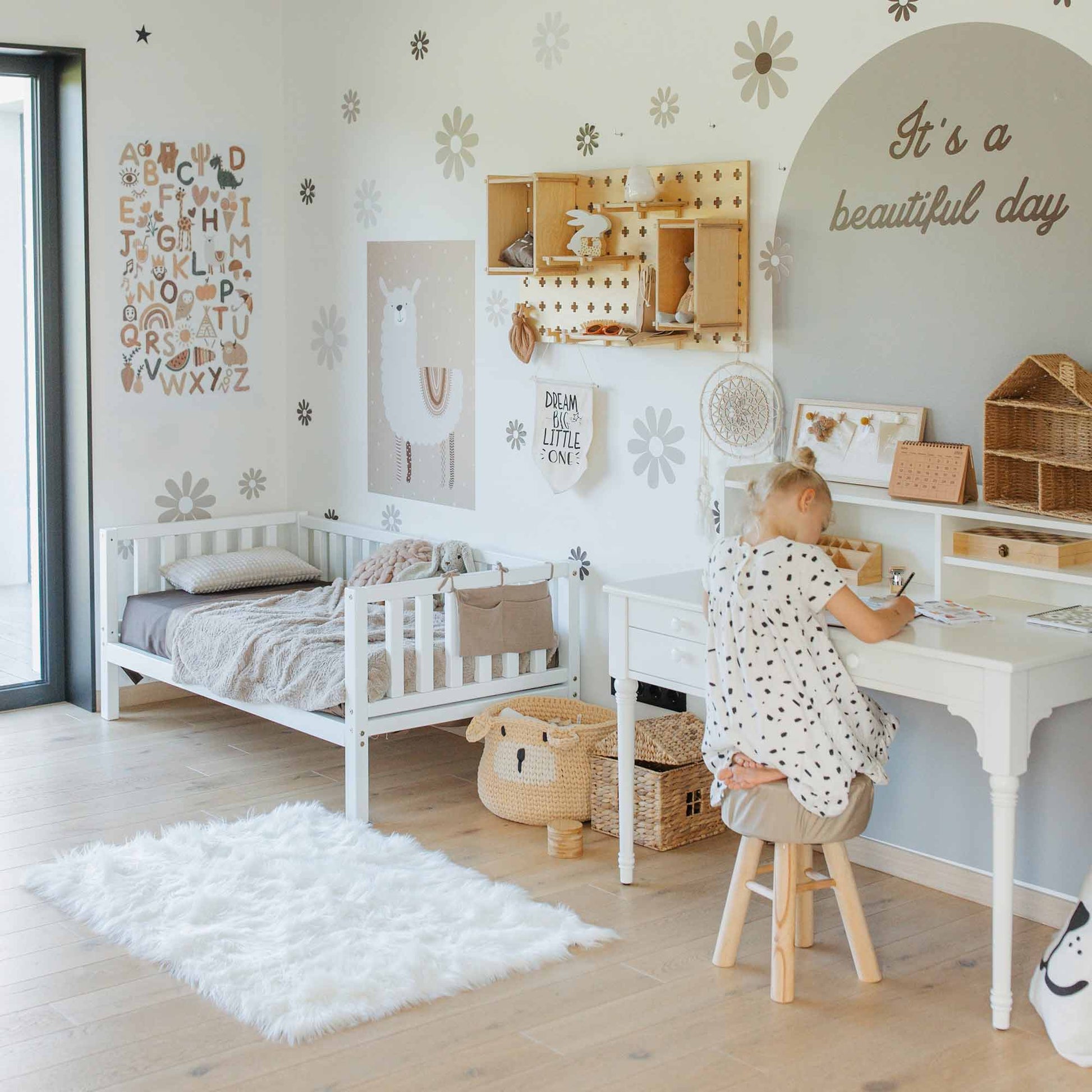 A child sits at a desk drawing in a bedroom with minimalist decor featuring neutral tones. The room includes a Montessori-inspired Kids' platform bed on legs with 3-sided rails and an "It's a beautiful day" wall decal.