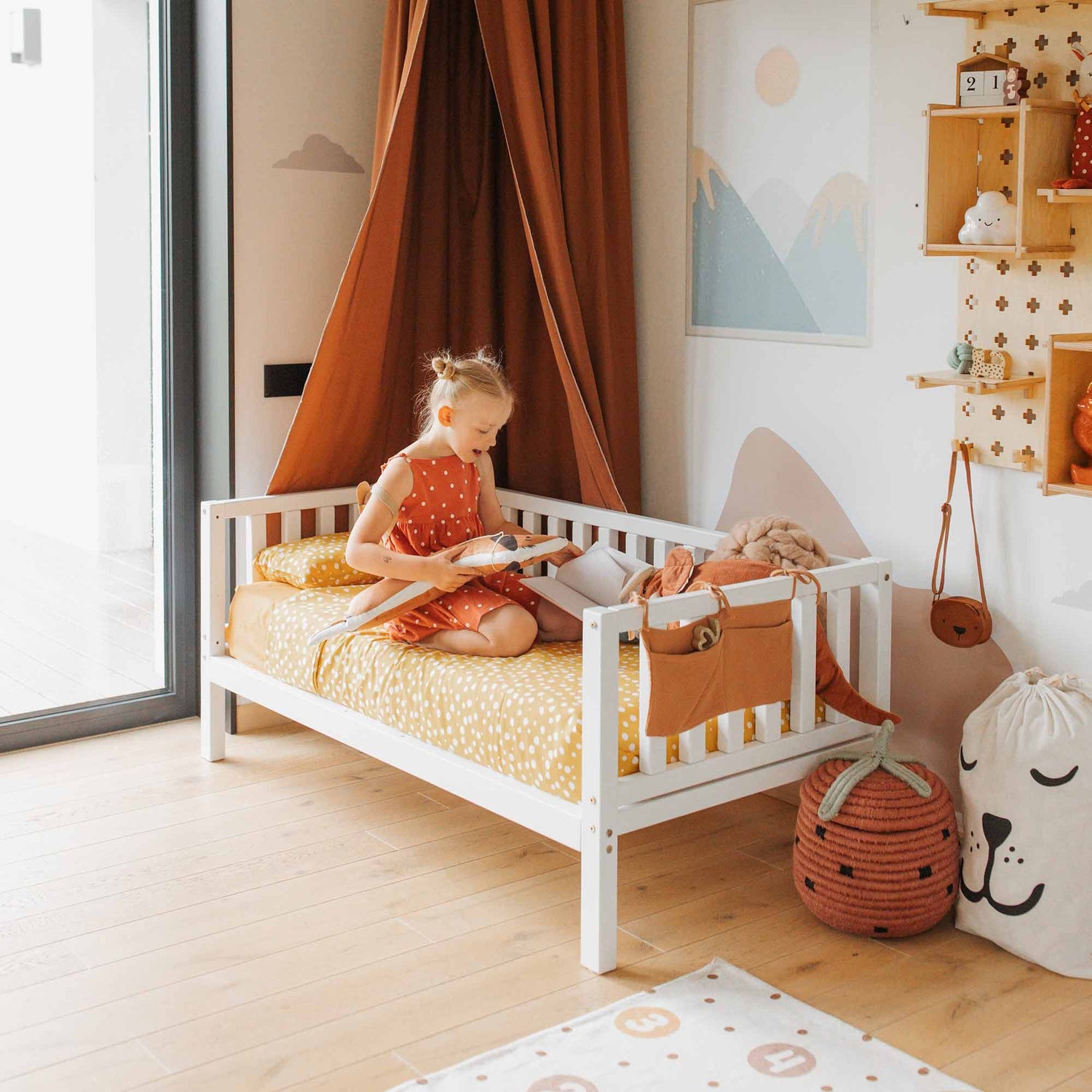 A young child sits on a cozy 2-in-1 toddler bed on legs with a 3-sided vertical rail in a neatly decorated bedroom, reading a book. The room features a wall with mountain artwork, a large window, and various decorative items.
