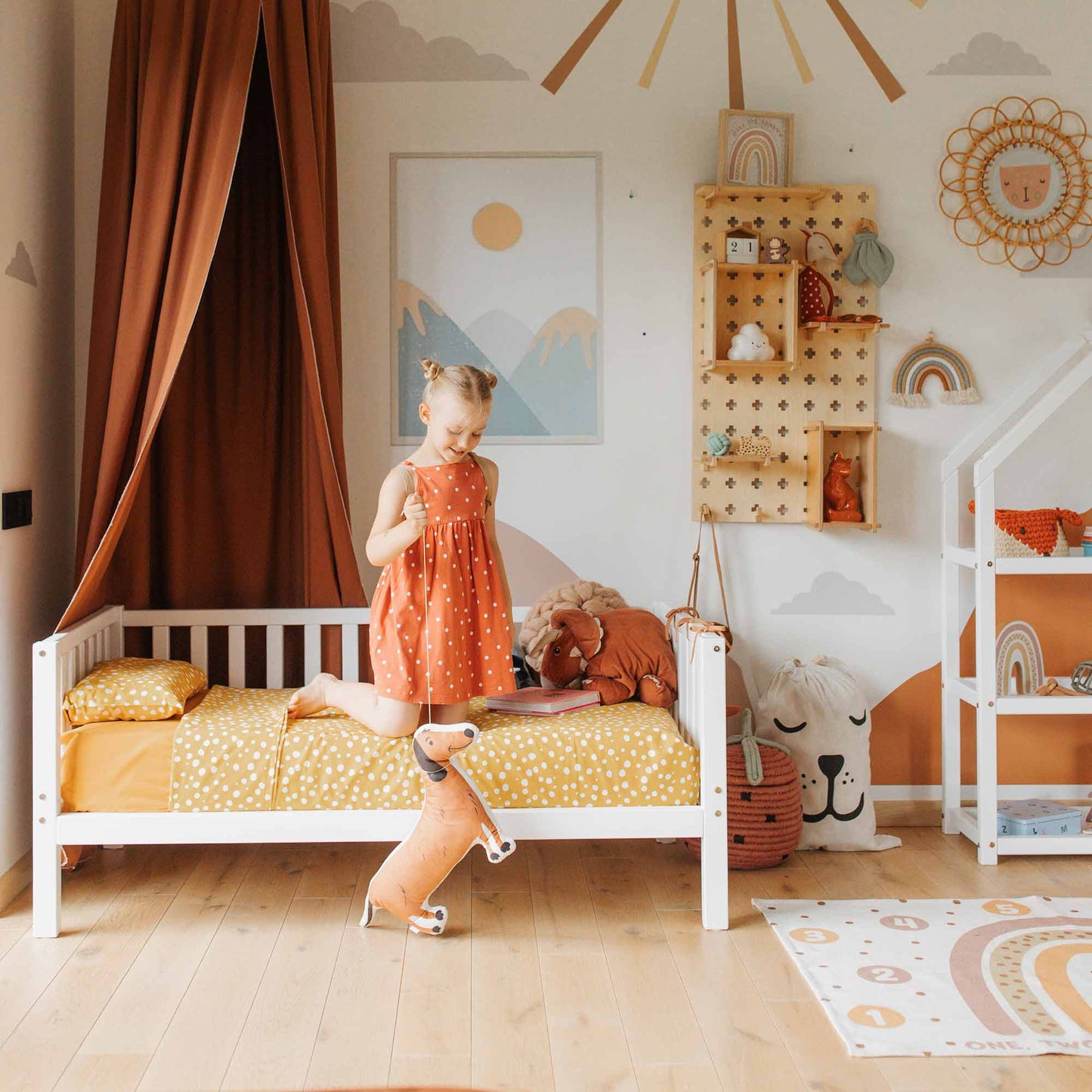 A young girl stands on her bed in a children's bedroom decorated with orange and white tones, including wall art, a canopy, and various stuffed animals. The Kids' platform bed on legs with 3-sided rails features sturdy wood legs, adding both stability and style to the Montessori-inspired room.