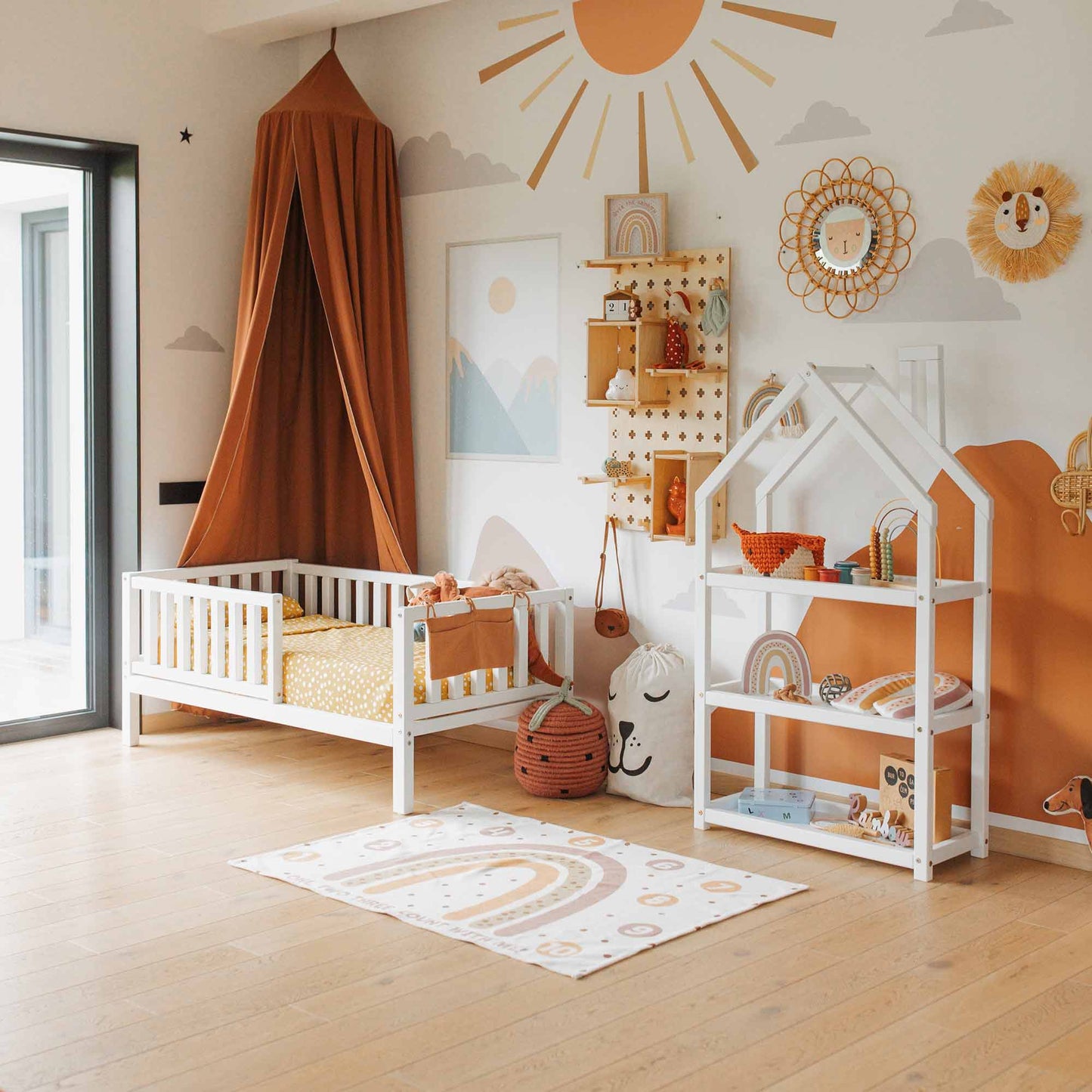 A children's room featuring a toddler bed on legs with a fence, canopy, house-shaped shelf unit, and pastel-colored decor. The walls are adorned with sun and rainbow murals to encourage independent sleeping, while various toys and storage items are neatly arranged.