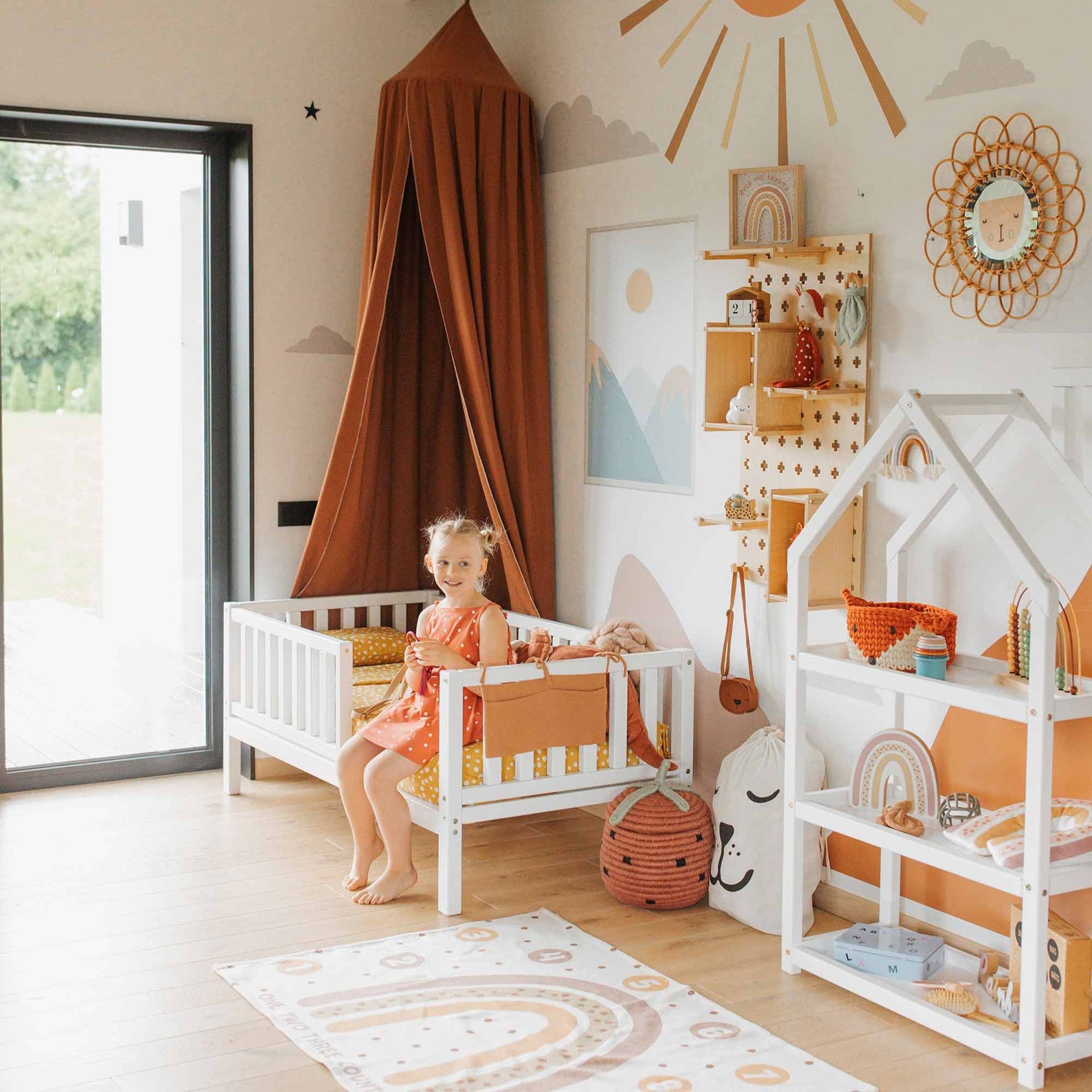 A young girl sits on a 2-in-1 toddler bed on legs with a vertical rail fence in a brightly decorated children's room featuring orange accents, rainbows, and sun-themed decor. A rug with a rainbow pattern lies on the wooden floor.