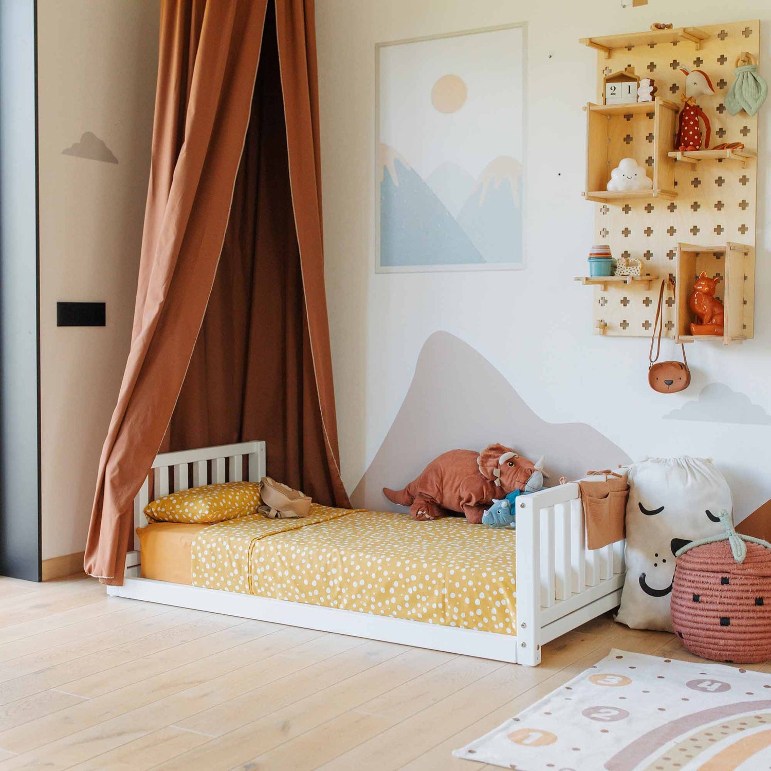 A child's bedroom featuring a Montessori-inspired kids' bed with a headboard and footboard, adorned with yellow bedding, surrounded by toys and storage baskets. A hanging curtain forms a canopy beside wall-mounted shelves holding various items.