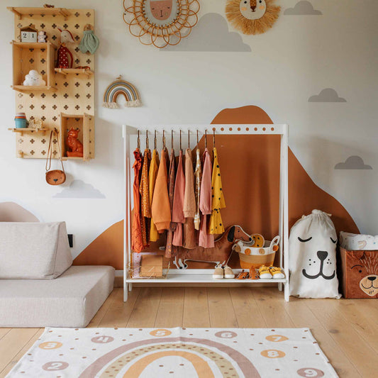 A children's room featuring a Montessori clothing rack adorned with colorful clothes, animal-themed decor, a wooden shelf filled with toys, a white sofa, and a rainbow-patterned rug on the floor.