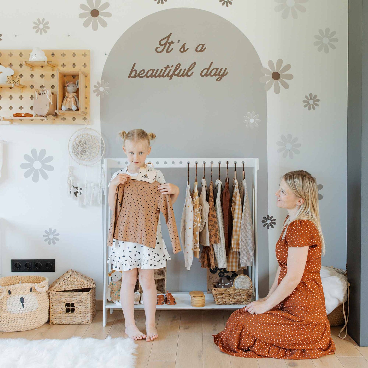A child in polka dot clothes holds up a brown shirt while an adult kneels nearby in a room decorated with flowers and the text "It's a beautiful day." Various clothes hang on a Montessori clothing rack, showcasing perfect closet organization.
