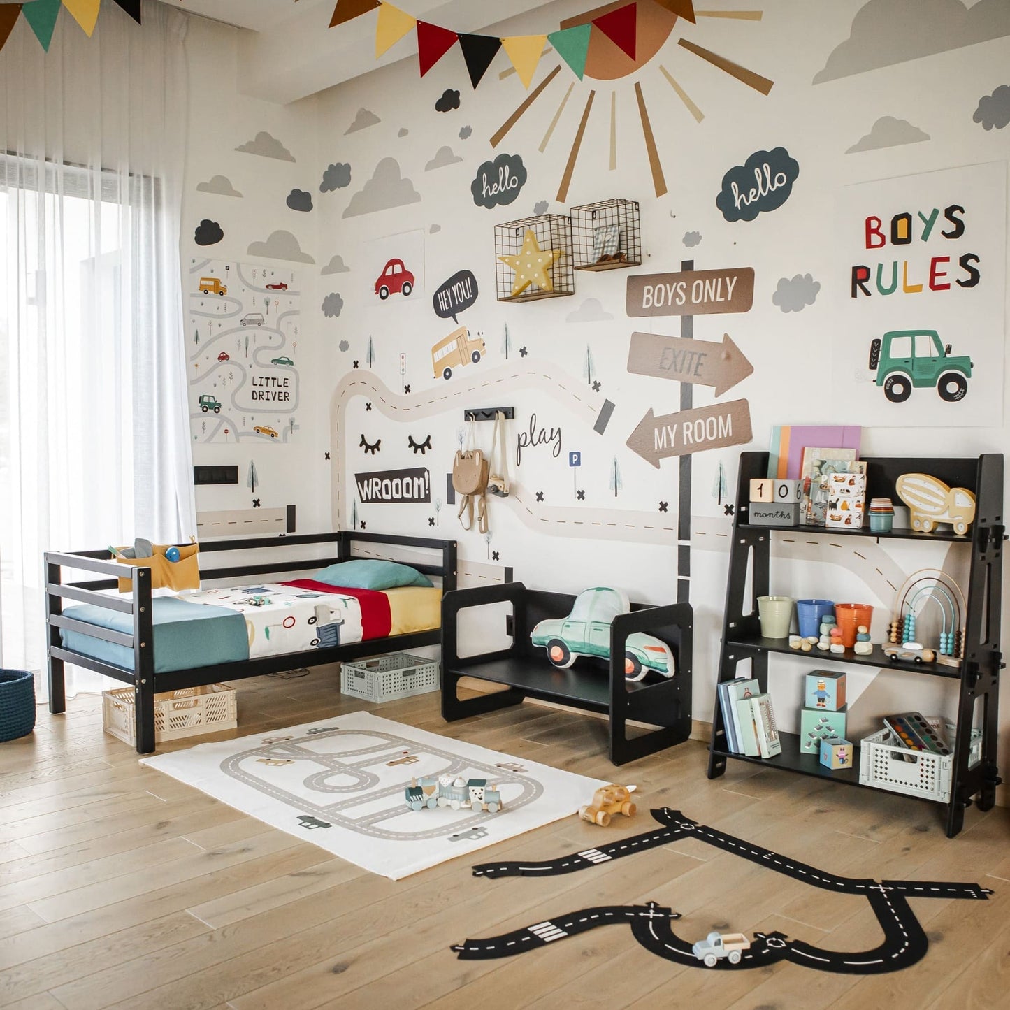 A car-themed kids' bedroom featuring a solid wood Kids' bed on legs with horizontal rail on 3-sides, shelves for toys and books, colorful wall décor, and a play rug outlined with roads and toy cars.