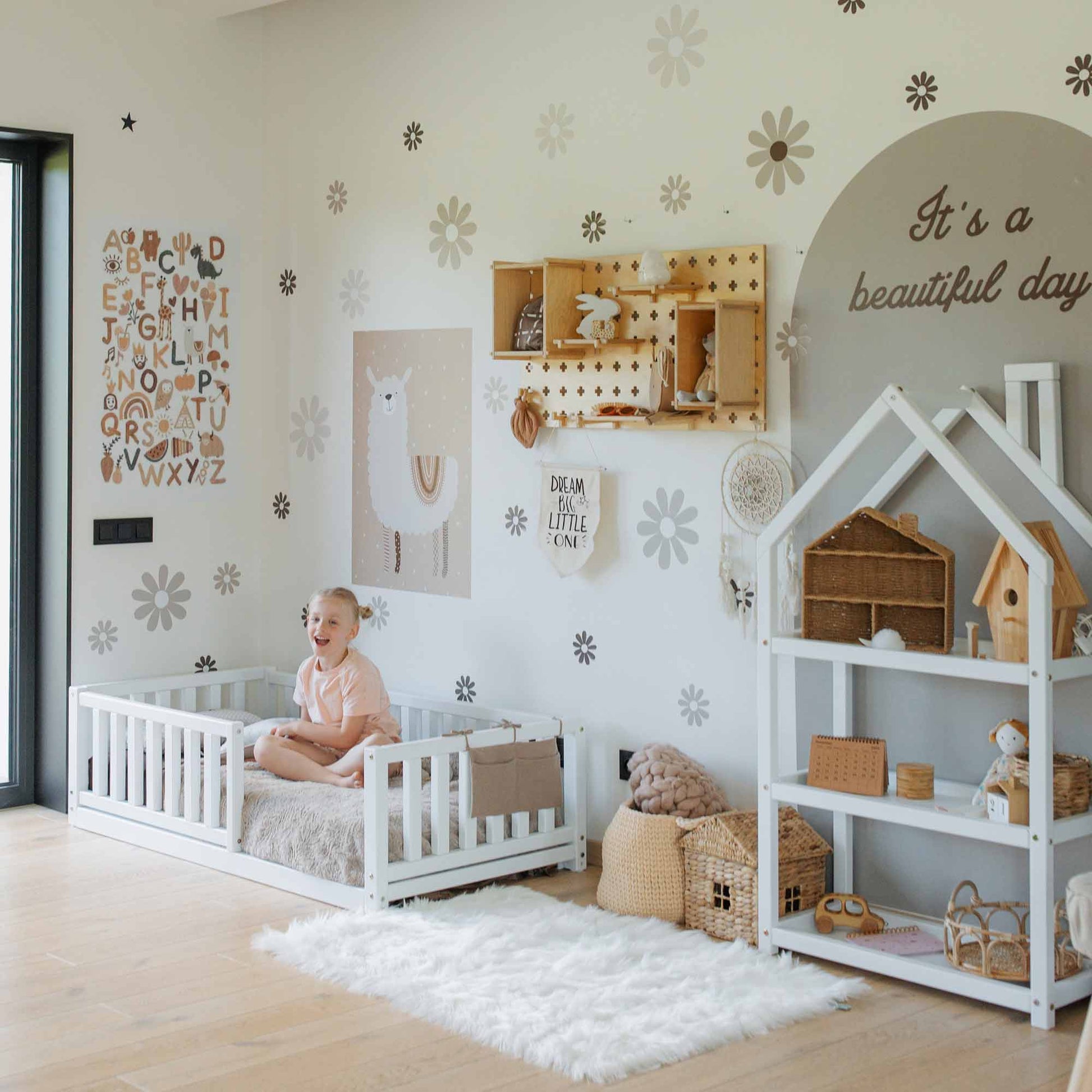 A child sits on a Montessori kids' bed with a fence in a well-decorated room adorned with wall art, shelves brimming with toys, and a fluffy white rug on the wooden floor, creating the perfect environment to encourage independent sleeping.
