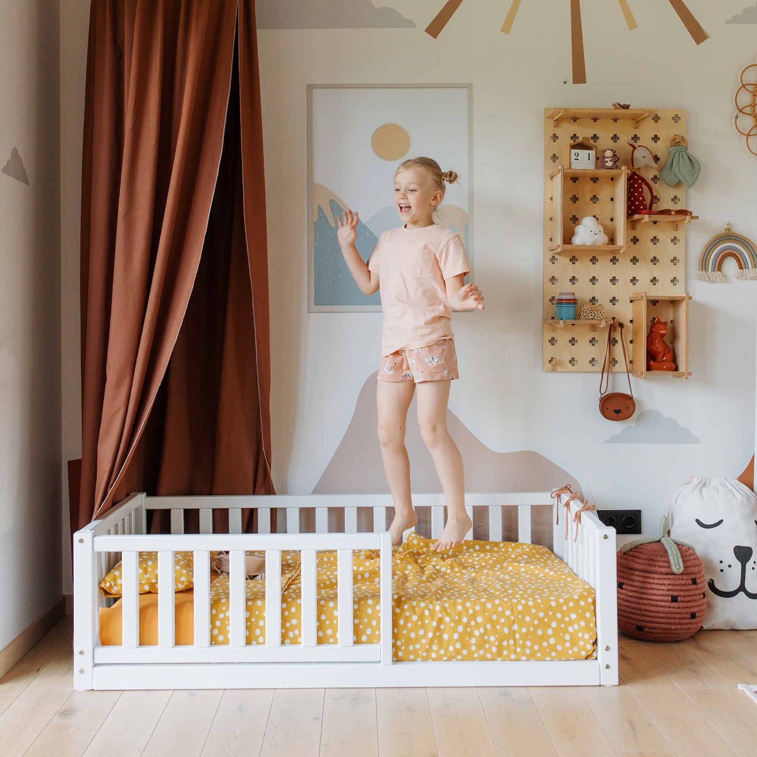 A child is standing and jumping on a Montessori kids' bed with a fence, adorned with orange and white polka dot bedding, in a brightly decorated room featuring brown curtains and various wall decorations, showcasing their excitement for independent sleeping.
