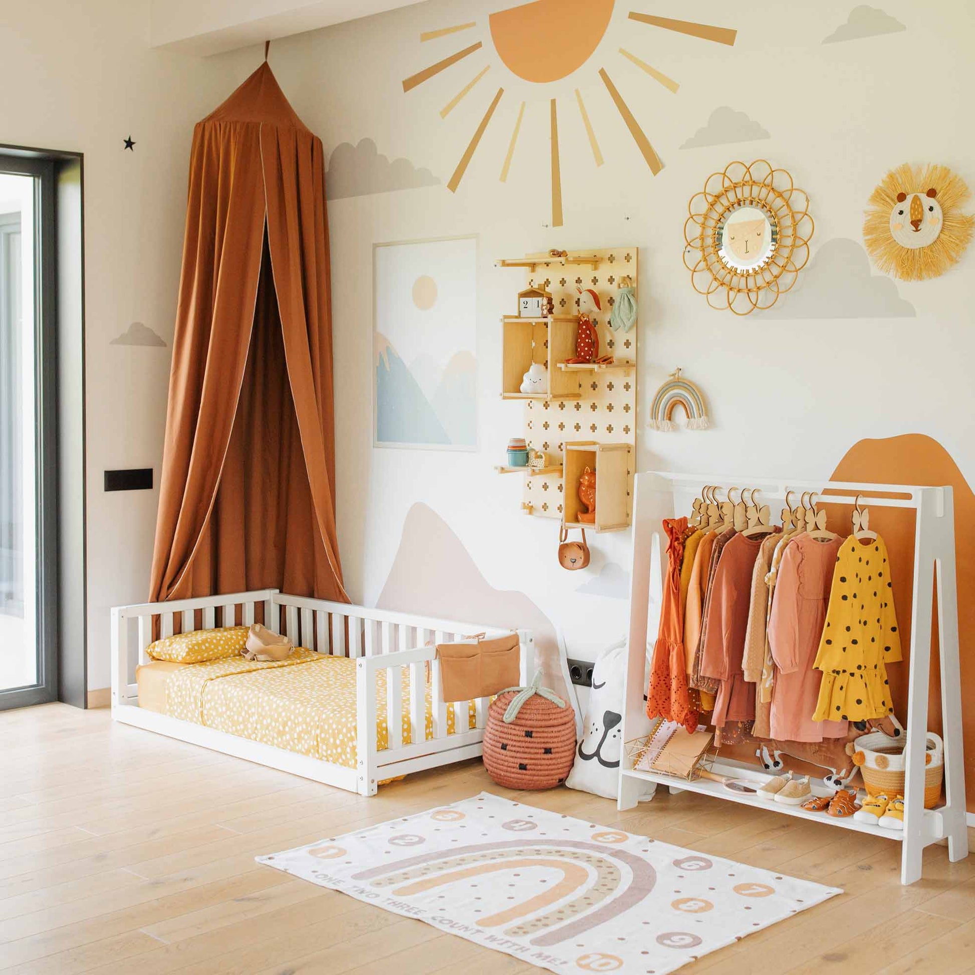 A brightly lit children's room featuring a white Montessori bed with 3-sided rails, an orange canopy, a rack of clothes, and decorative wall art in tones of orange, yellow, and white showcasing motifs of the sun, rainbow, and lion.