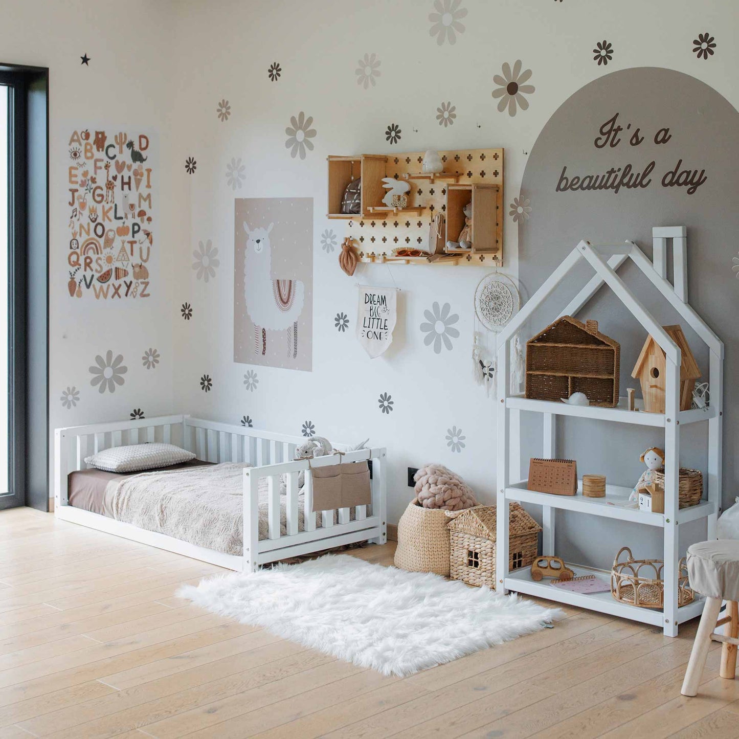 A child's bedroom with a Montessori bed featuring 3-sided rails, a playhouse shelf, and various wall decorations including alphabet and llama posters, wooden shelves, and an "It's a beautiful day" sign.