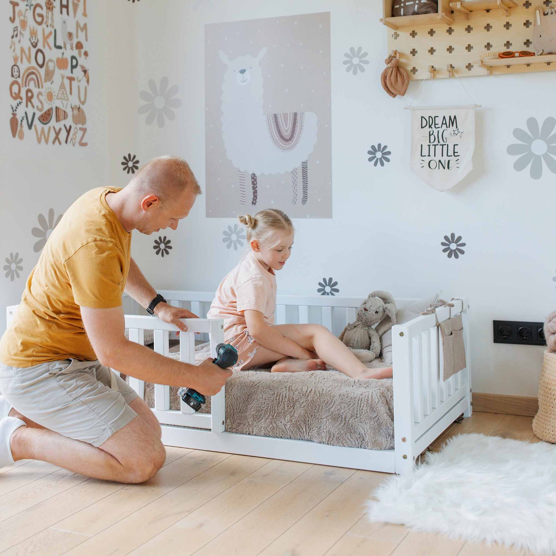 A man in an orange shirt uses an electric drill while assembling the Montessori kids' bed with a fence in a child's room. A little girl sits on the bed, which is decorated with grey blankets and plush toys, eagerly awaiting her new space for independent sleeping.