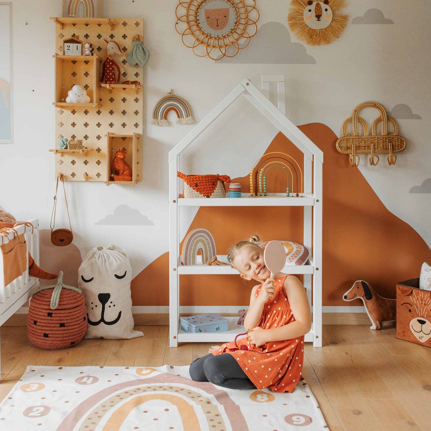 A young child in an orange dress sits on the floor of a children’s room holding a toy. The space is decorated with colorful rainbow-themed items and stuffed animals displayed on charming House-shaped Montessori shelves.