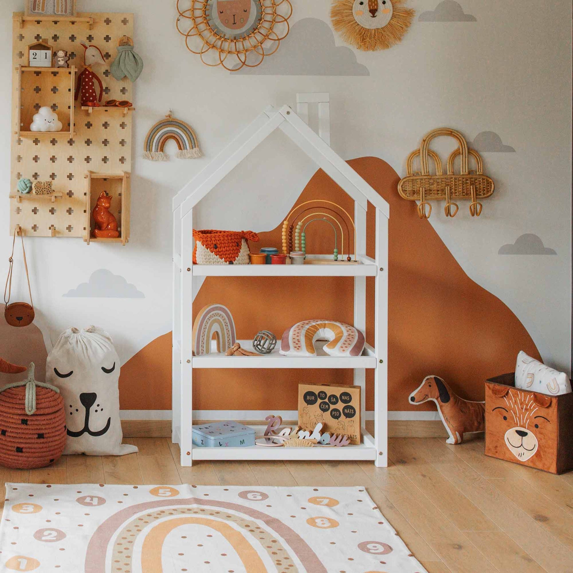 A child's room with a House-shaped Montessori shelf, toys, and decor in earthy tones. A wall painted with clouds and rainbows, and a playmat featuring a rainbow pattern are visible.