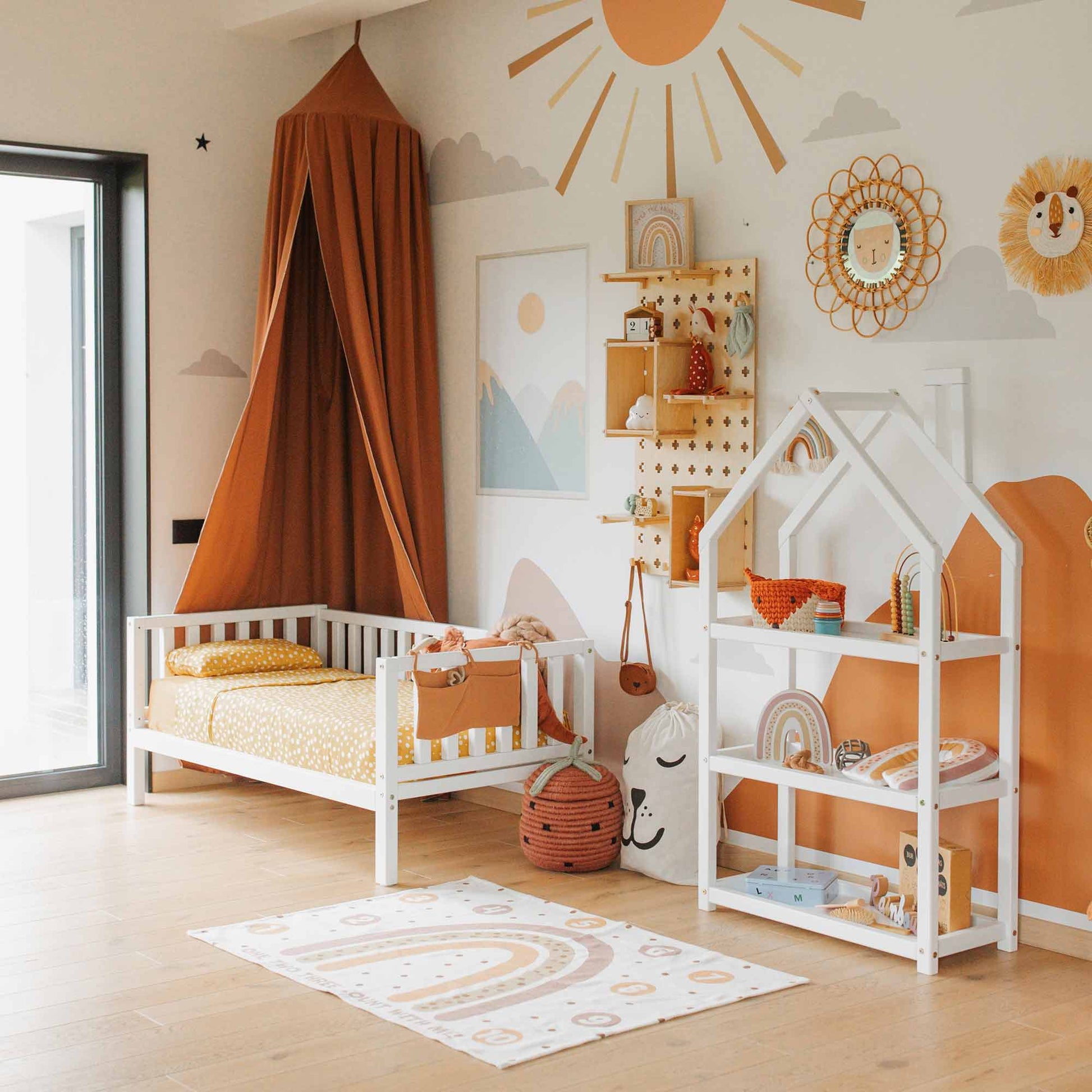 A children's room with a bed, a canopy, a House-shaped Montessori shelf, wall decorations, and a play rug. Decor includes sun and rainbow motifs in warm tones. A glass door is on the left side.